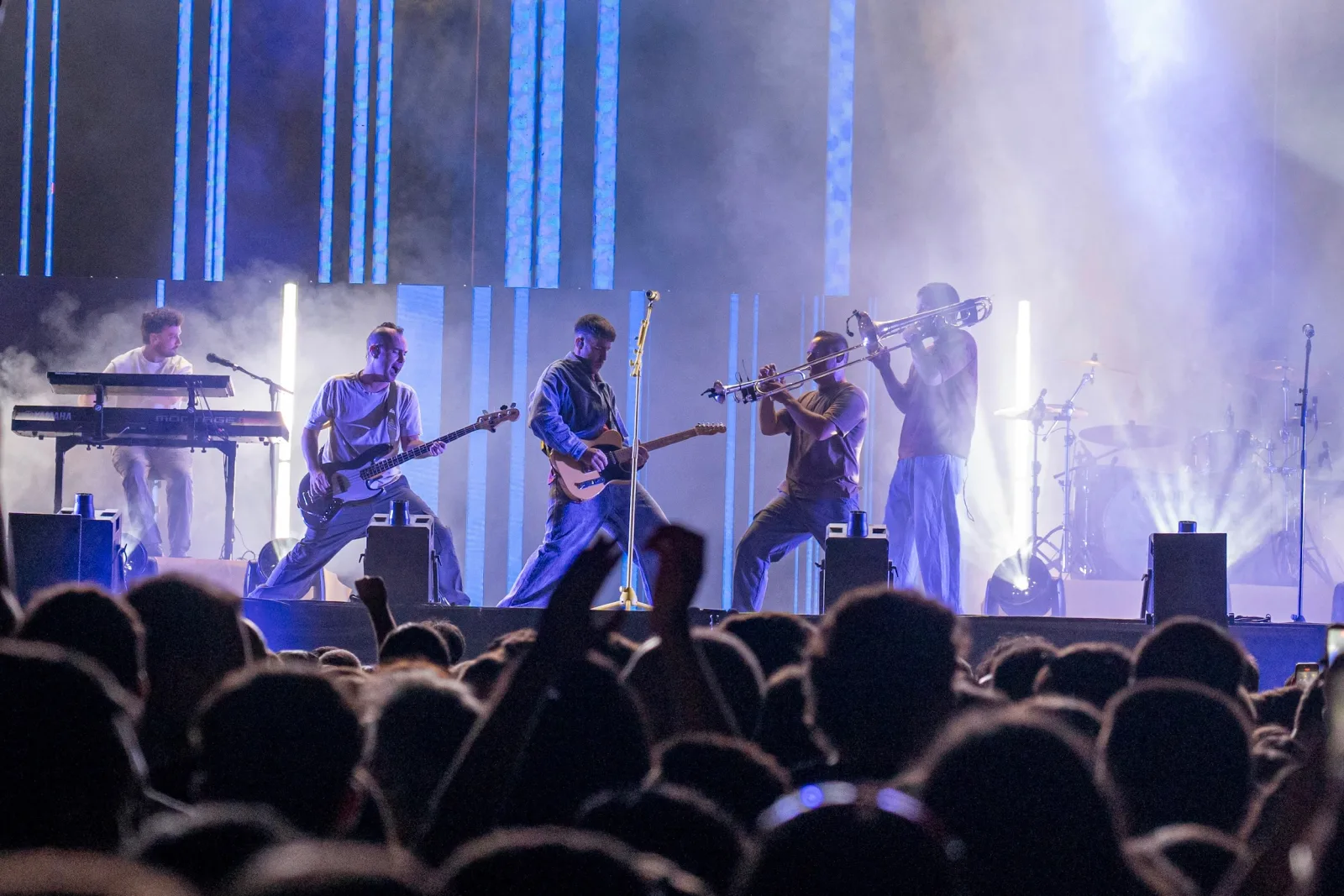 Cinco músicos sobre el escenario —teclado, bajo, guitarra, trompeta y trombón— bajo luces azules, con las cabezas del público en primer término