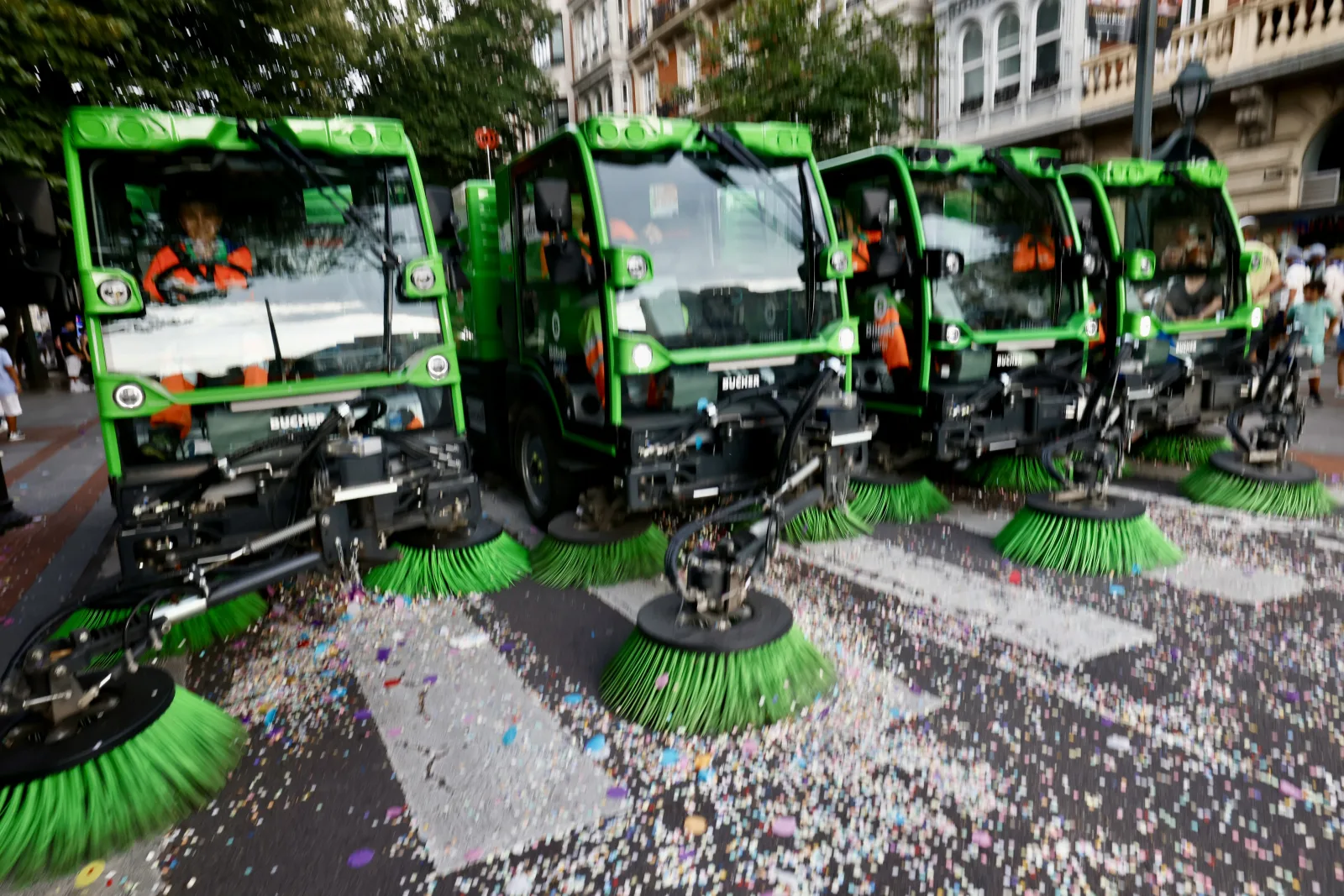 Cuatro barredoras verdes alineadas en diagonal avanzan con sus cepillos girando sobre una calzada cubierta de confeti de colores