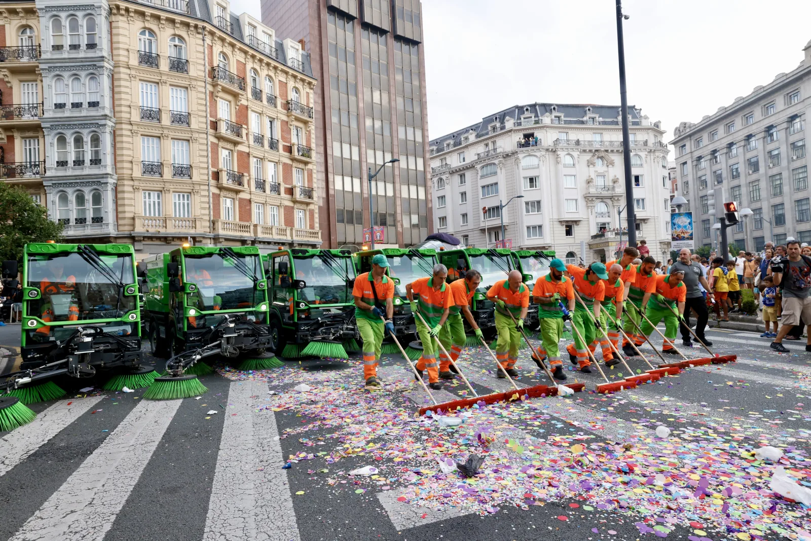 Vista frontal de una fila de operarios empujando escobones rojos sobre una calzada llena de confeti, con cuatro barredoras verdes aparcadas a la izquierda y edificios señoriales al fondo