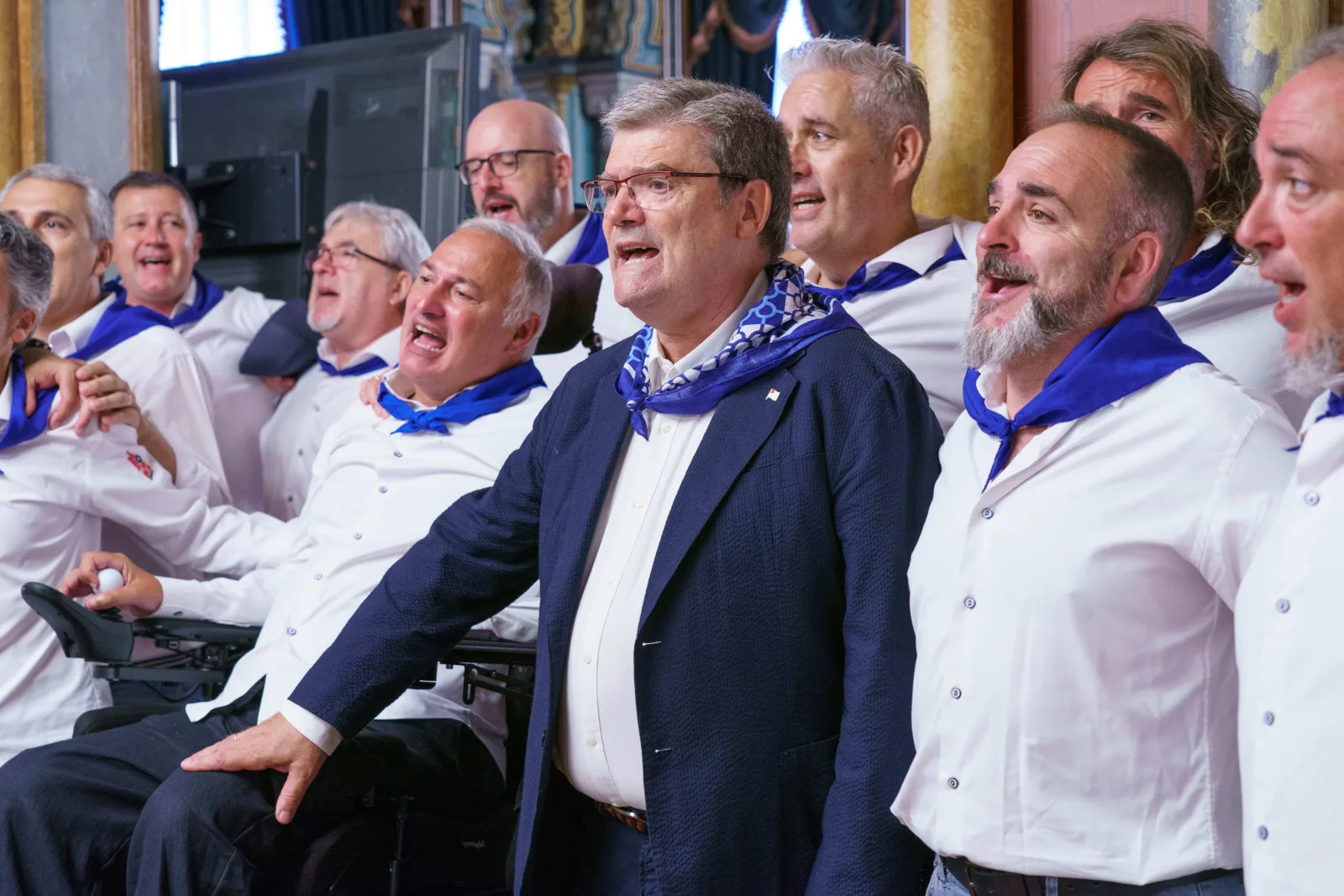 Un grupo de cantores con camisa blanca y pañuelo azul canta junto al Alcalde de Bilbao dentro del salón