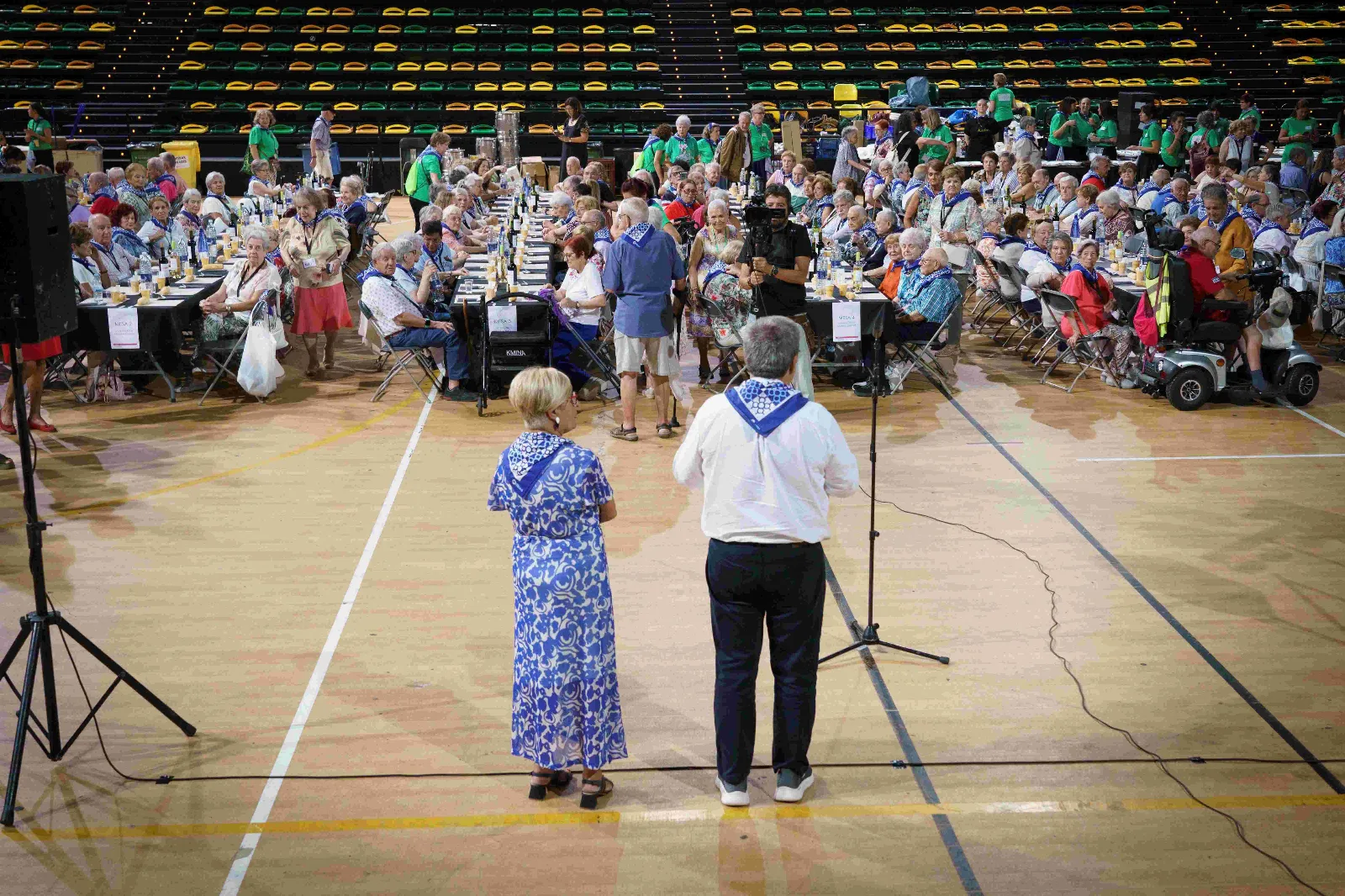 Vista general del pabellón del Bilbao Arena con largas mesas ocupadas por cientos de comensales con pañuelo azul y las gradas al fondo; en primer plano, dos personas de espaldas ante un micrófono
