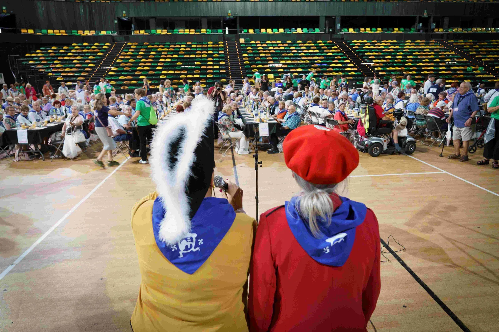Dos personas de espaldas con pañuelo azul, una con chaqueta amarilla y tocado de plumas y otra con chaqueta y txapela rojas, ante el pabellón lleno de comensales