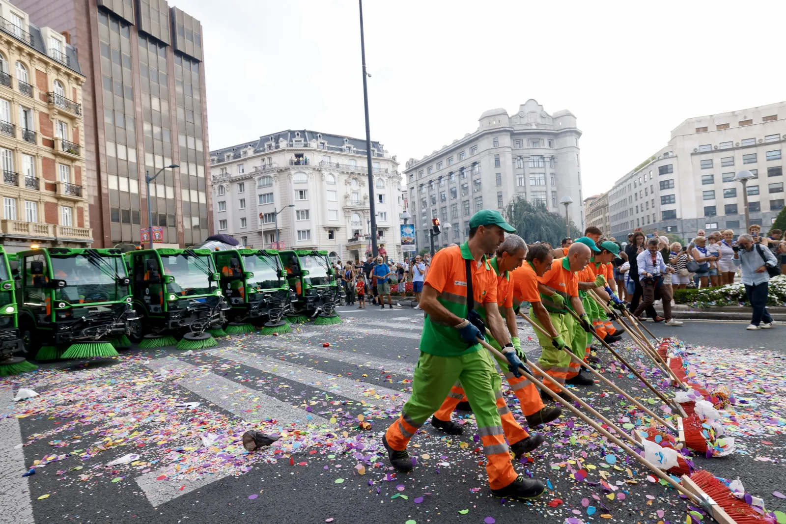 Una fila de operarios con escobones rojos avanza por una avenida ancha cubierta de confeti mientras cuatro barredoras esperan a su izquierda y varias personas miran desde la acera