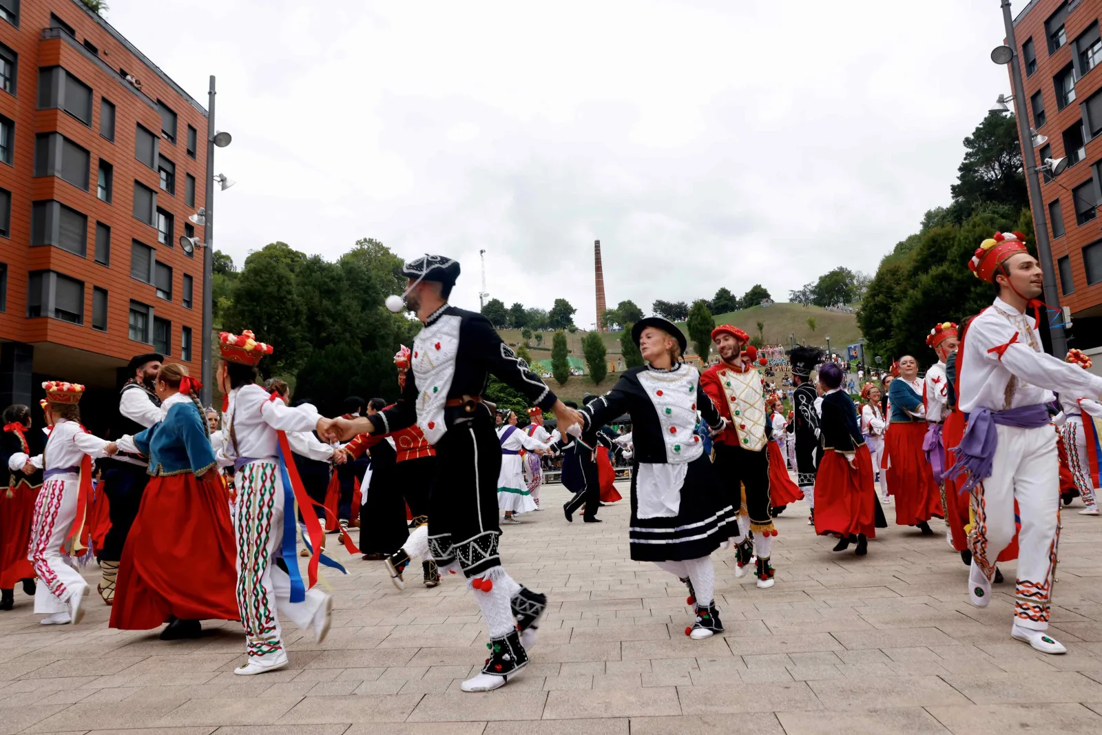 Un corro abierto de dantzaris con traje tradicional recorre la plaza cogidos de la mano, con los bloques de ladrillo detrás