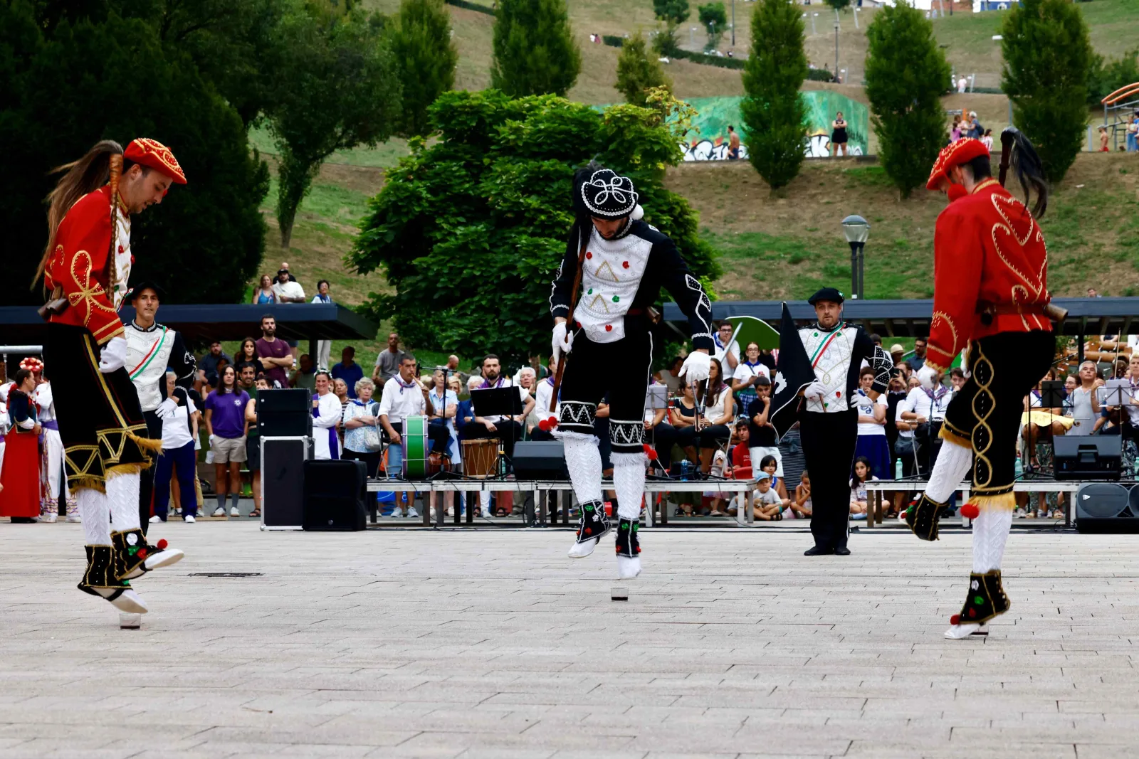 Tres dantzaris de rojo y negro saltan sobre el pavimento de la plaza ante el público, con un músico al fondo