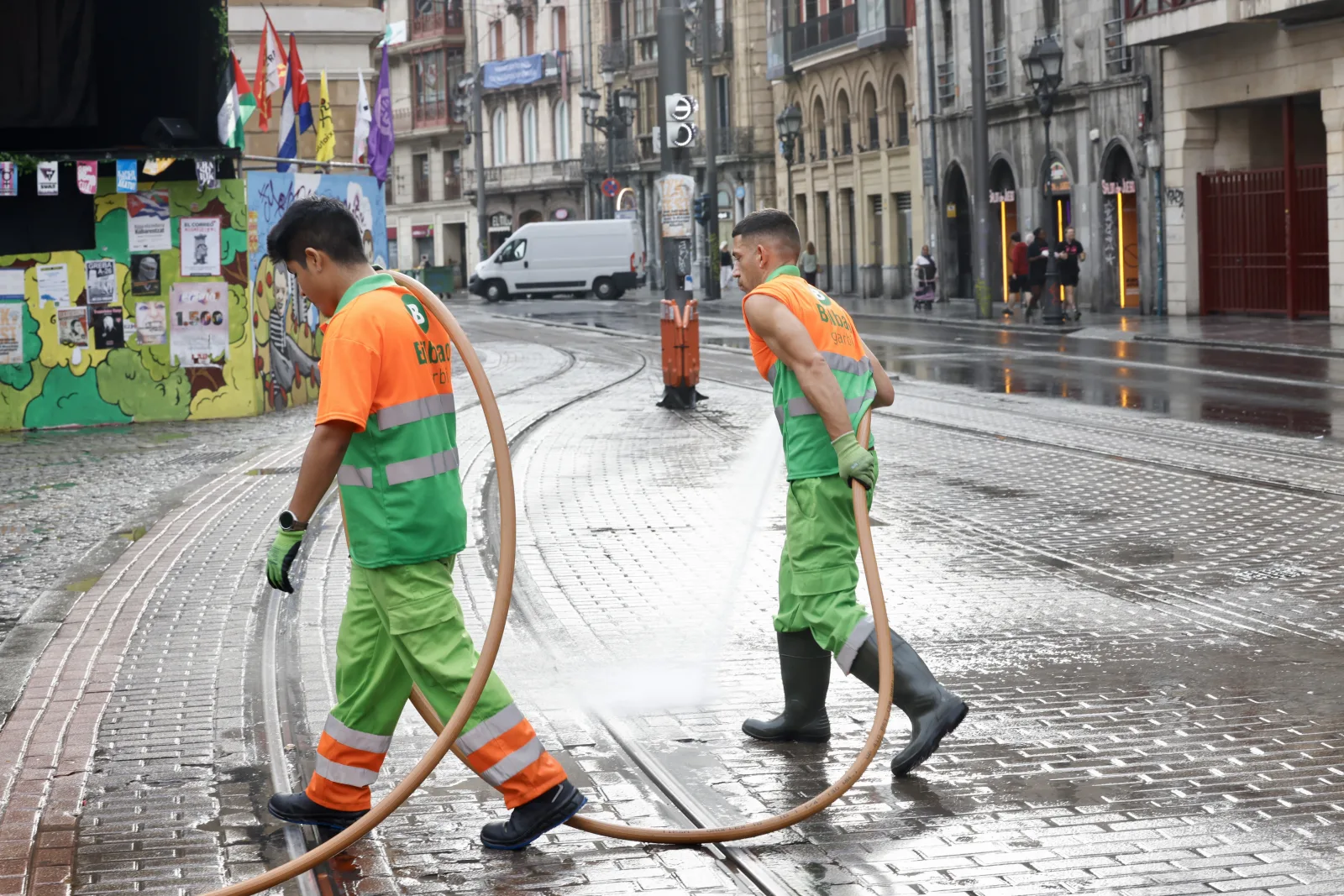 Dos operarios con uniforme naranja y verde arrastran una manguera larga cruzando una calle mojada con vías de tranvía, con la ciudad casi vacía al amanecer
