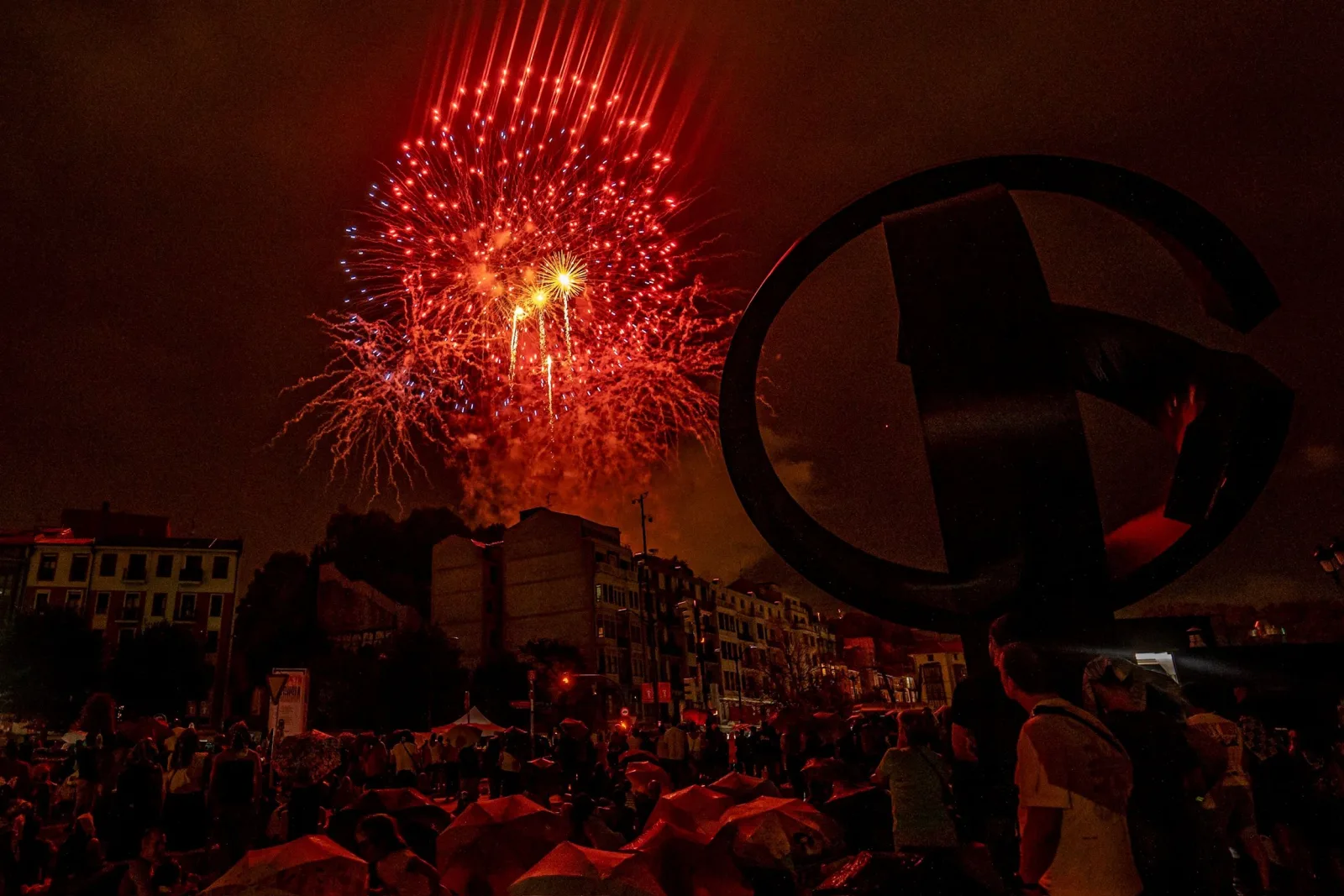 Un gran estallido rojo de fuegos artificiales sobre los edificios de la ría de Bilbao tiñe de rojo los paraguas del público