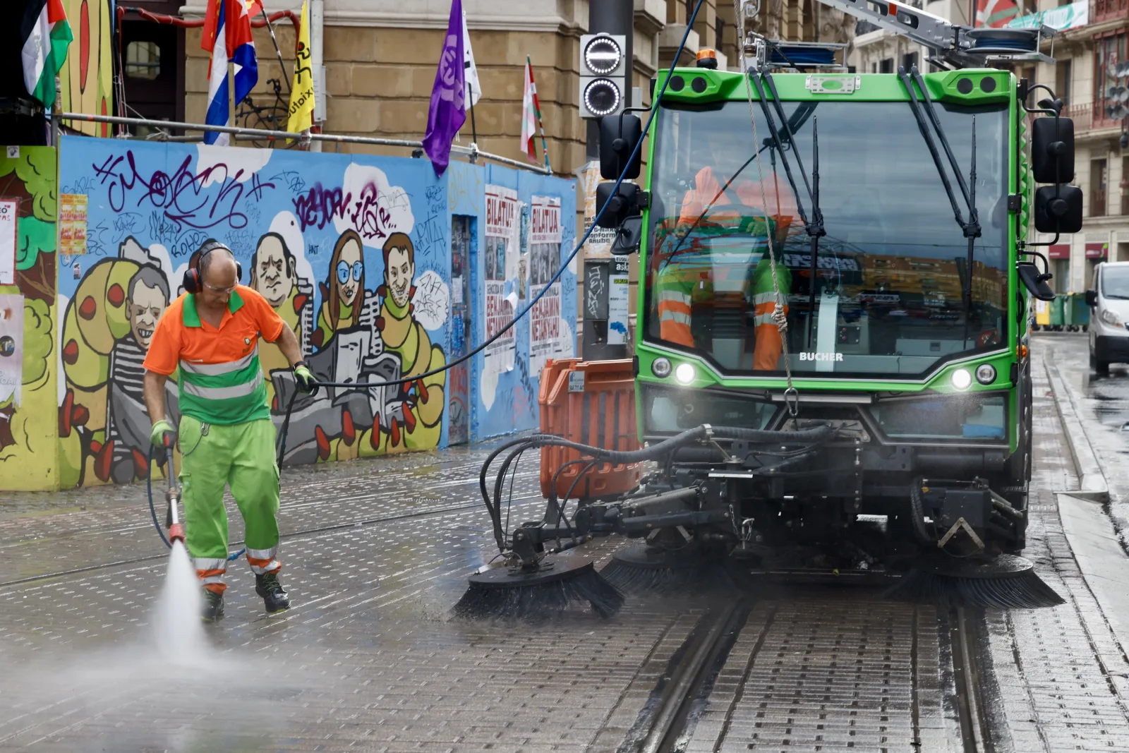 Un operario con uniforme naranja y verde y cascos de protección auditiva baldea con una lanza de agua a presión junto a una barredora verde, con un mural pintado y vías de tranvía al fondo