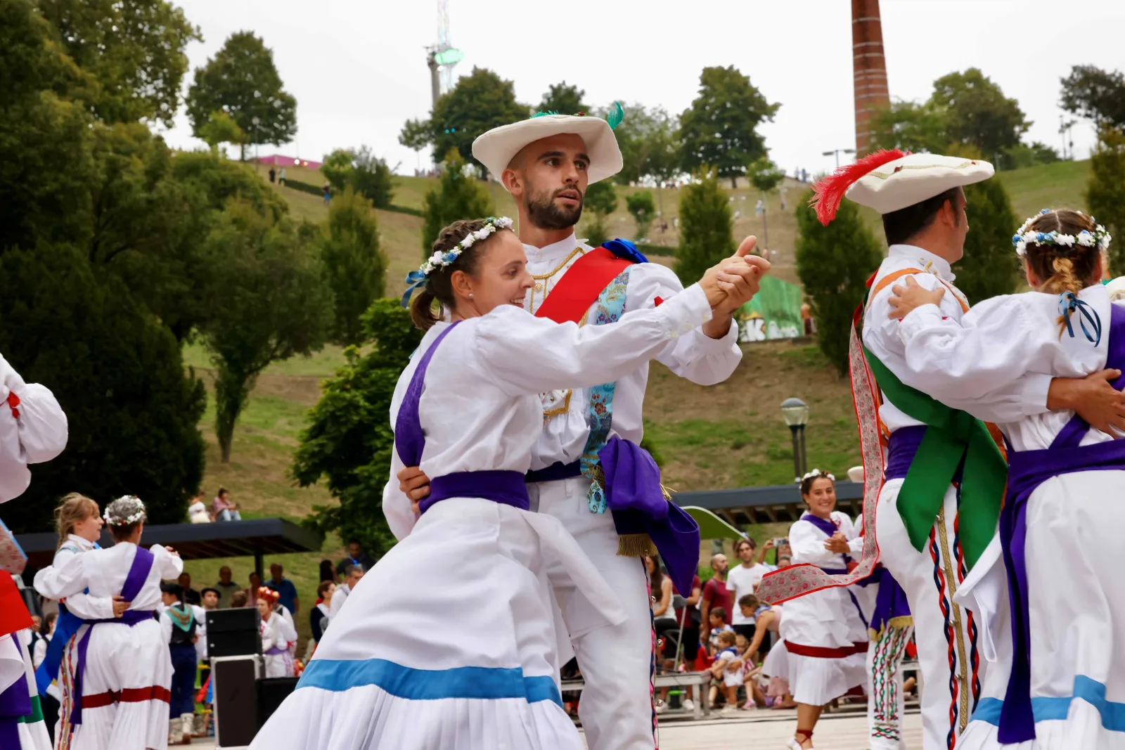 Una pareja de dantzaris baila agarrada, con más parejas al fondo y la ladera verde del parque detrás