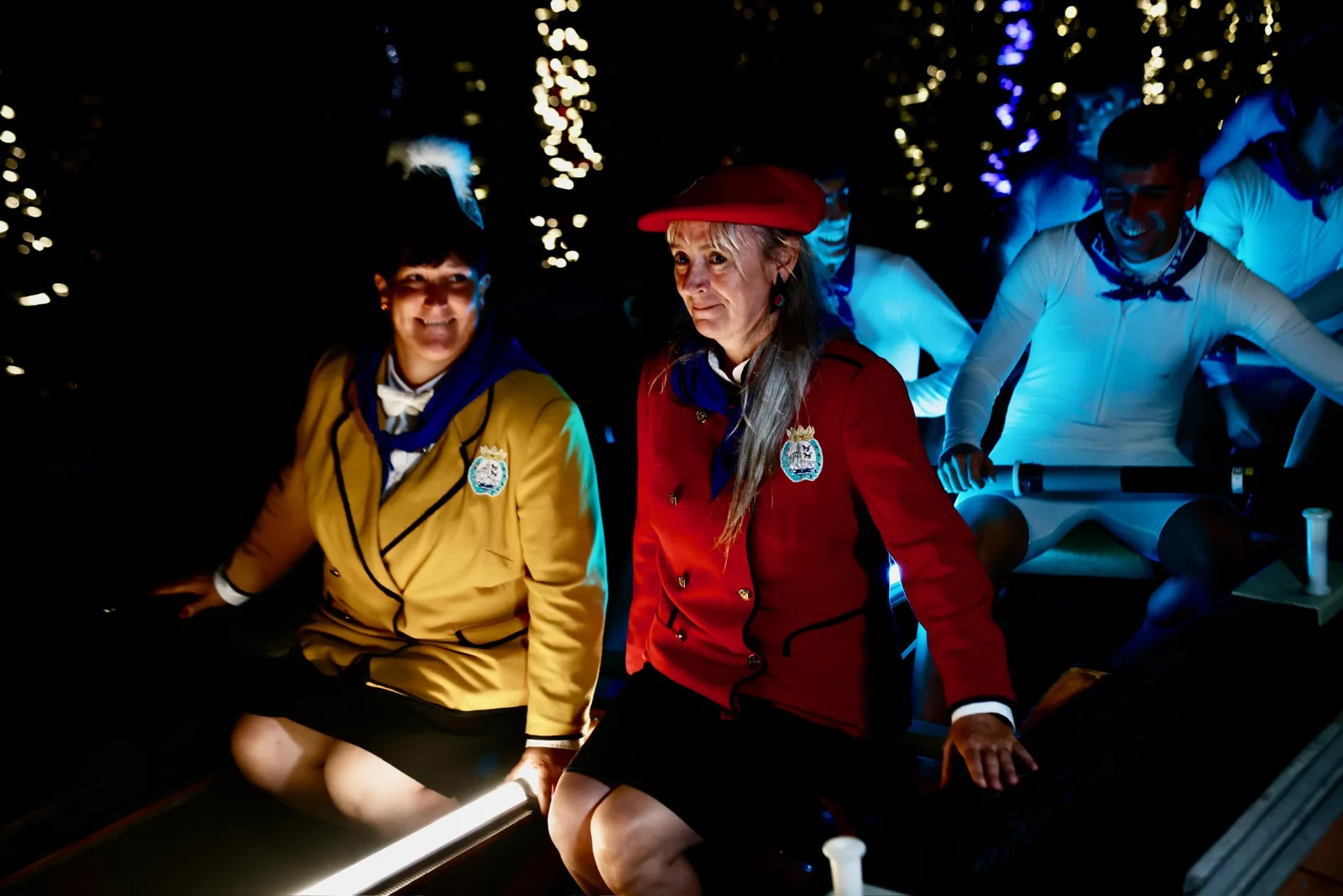 Dos mujeres con las chaquetas de gala amarilla y roja del Ayuntamiento de Bilbao viajan sentadas en una trainera iluminada, con remeras y remeros de blanco detrás y las luces de la ría al fondo