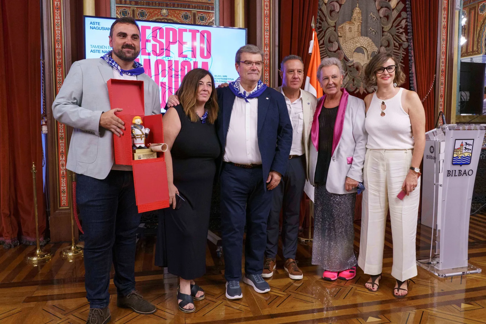 Un grupo posa con la figura del reconocimiento dentro de su caja roja abierta, con la imagen de la campaña Errespetu Nagusia en la pantalla del fondo