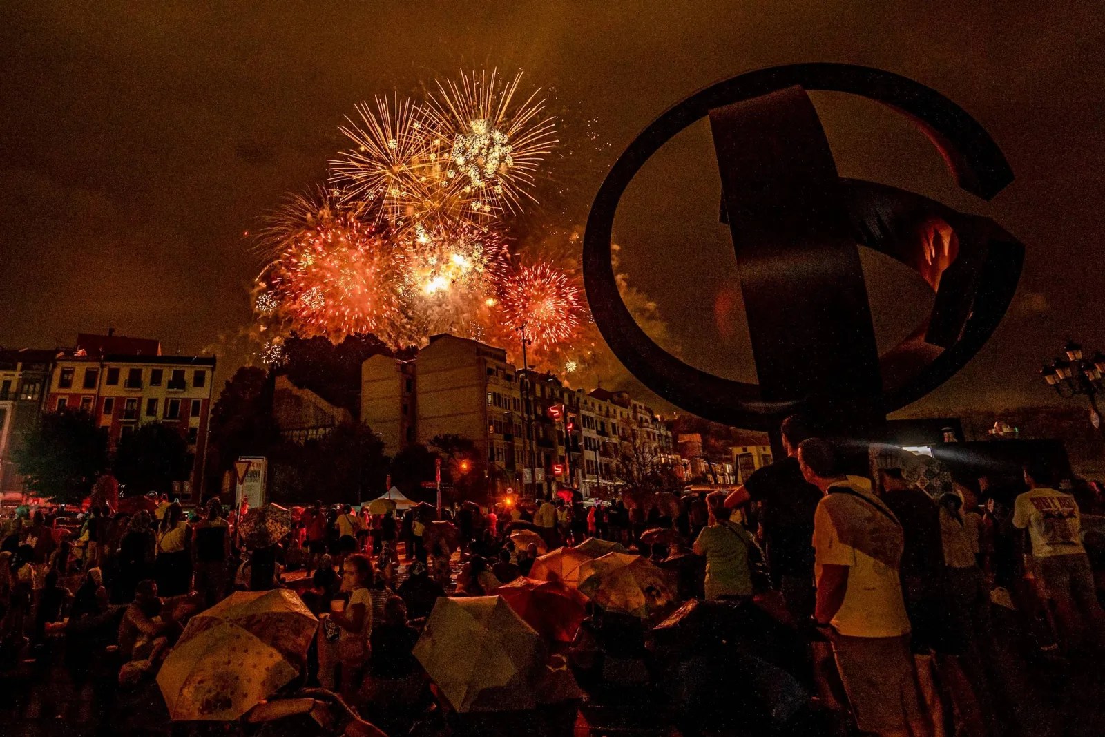 Decenas de personas, de pie y sentadas bajo paraguas, miran hacia varios fuegos artificiales naranjas y blancos que estallan sobre los edificios