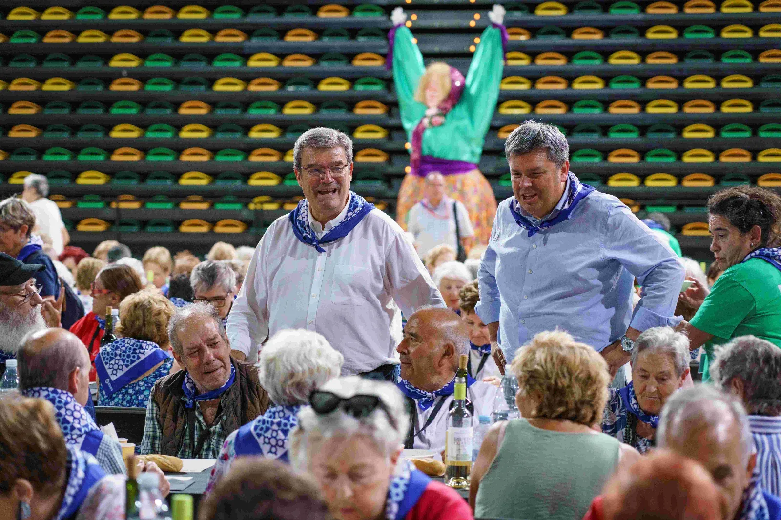 Dos representantes municipales con pañuelo azul conversan de pie entre las mesas, con Marijaia al fondo sobre las gradas