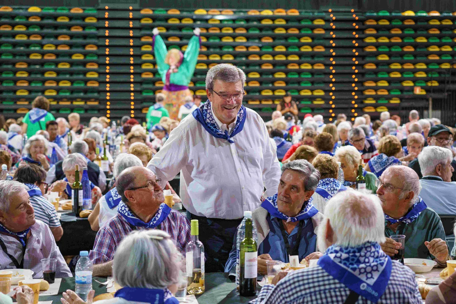 Un representante municipal con camisa blanca y pañuelo azul conversa de pie entre las mesas mientras los comensales comen; al fondo, Marijaia sobre las gradas