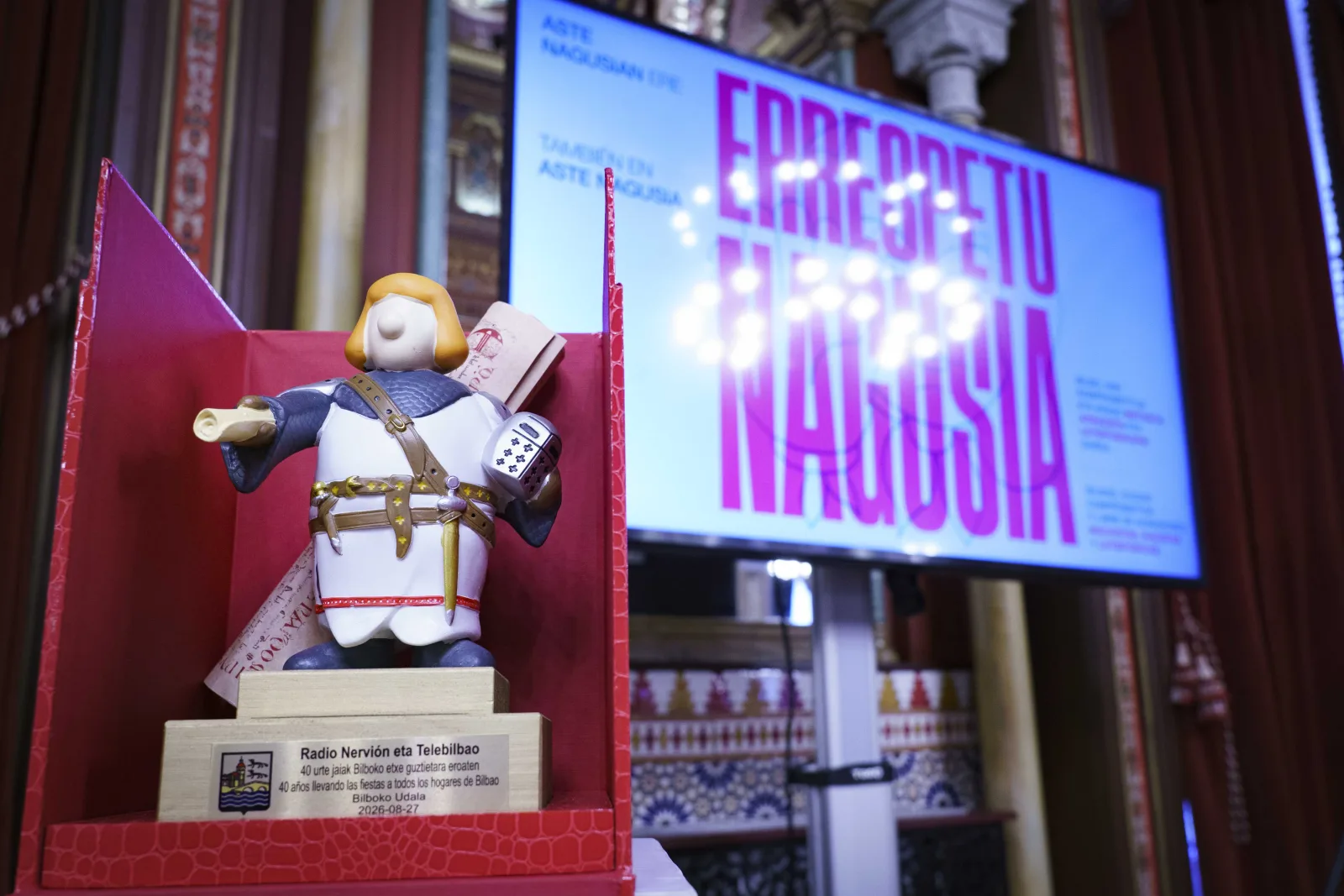 Primer plano de la figura del reconocimiento sobre su peana de madera, con la placa grabada a Radio Nervión eta Telebilbao y la fecha 2026-08-27