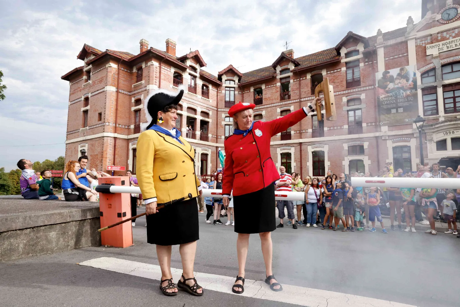 La Txupinera, de chaqueta amarilla, y la Pregonera, de chaqueta roja, saludan en el recinto del hospital con público al fondo