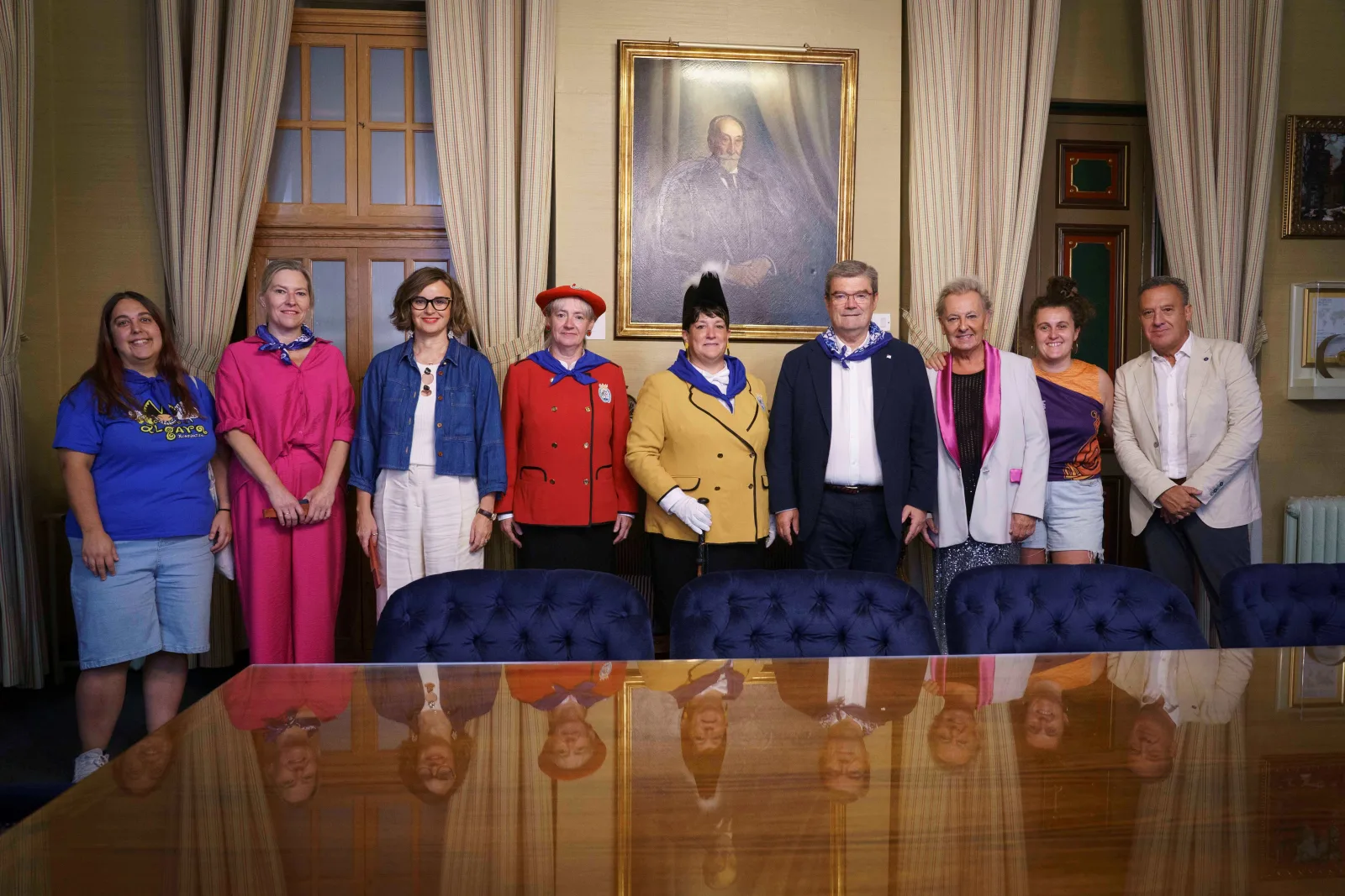 Nueve personas posan de pie tras una mesa de juntas del Ayuntamiento; entre ellas, la Txupinera de chaqueta amarilla y la Pregonera de chaqueta roja