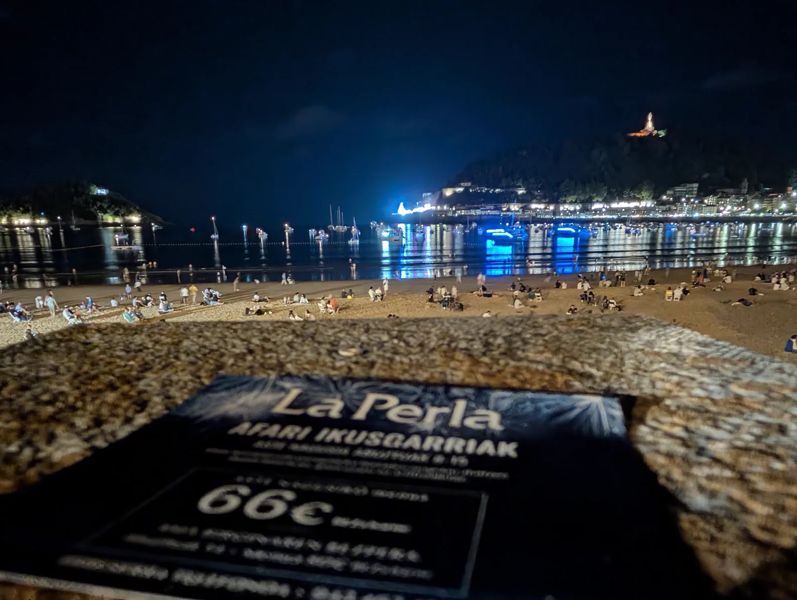 Cartel del Restaurante La Perla en la barandilla del paseo anunciando las cenas de Semana Grande por 66 euros, con la playa llena de gente al fondo
