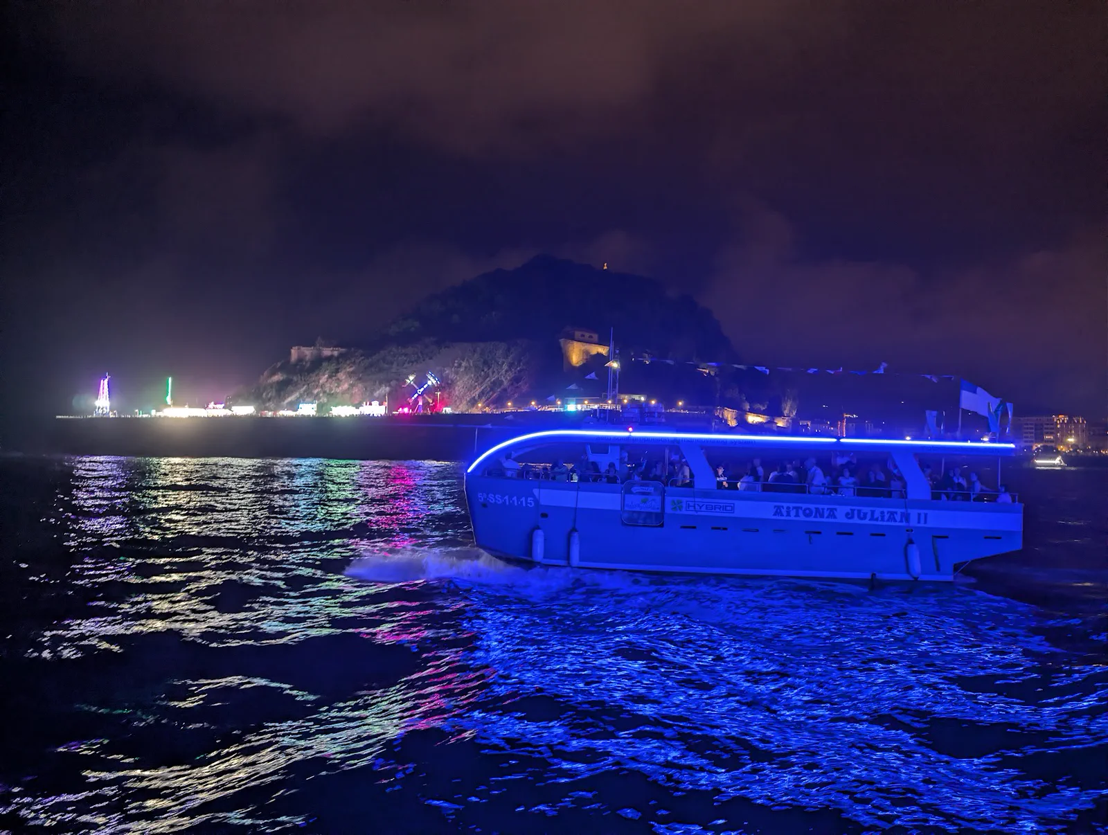 El Aitona Julián II navegando de noche de vuelta al puerto con su tira de LED azul encendida, con el monte Urgull y las luces de las ferias del Paseo Nuevo al fondo
