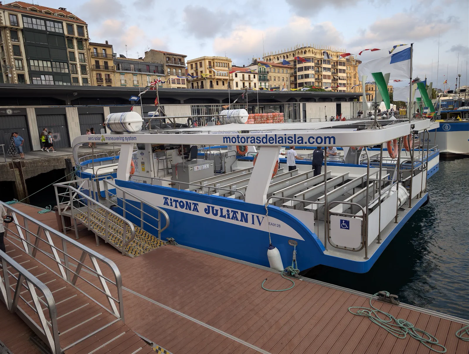 El Aitona Julián IV amarrado y vacío en el muelle de Donostia con la pasarela puesta, los bancos corridos a la vista y las casas de la Parte Vieja empavesadas al fondo