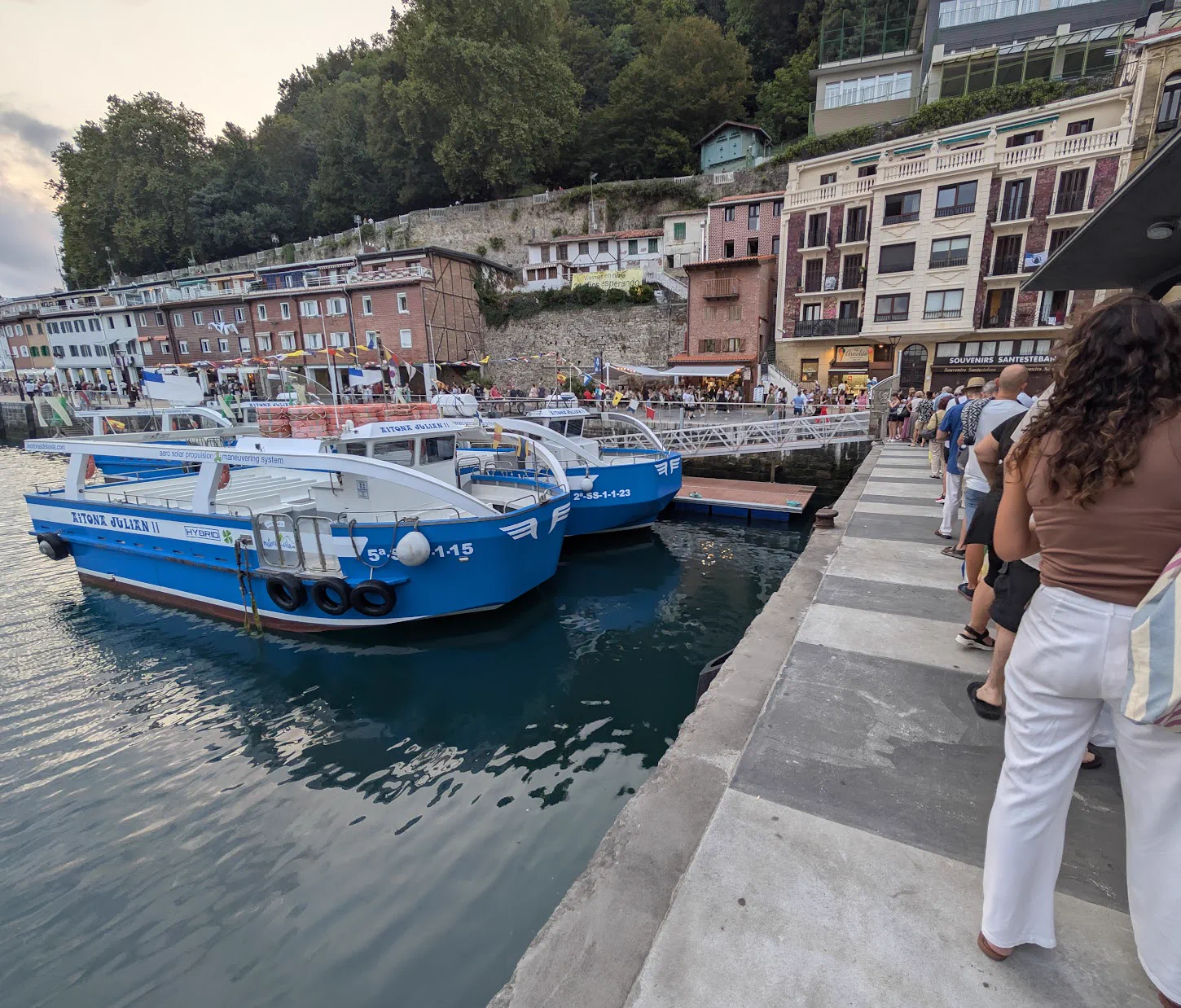 Cola de gente esperando en el muelle del puerto de Donostia junto a dos motoras azules amarradas, con la heladería Arnoldo y las casas del muelle al fondo