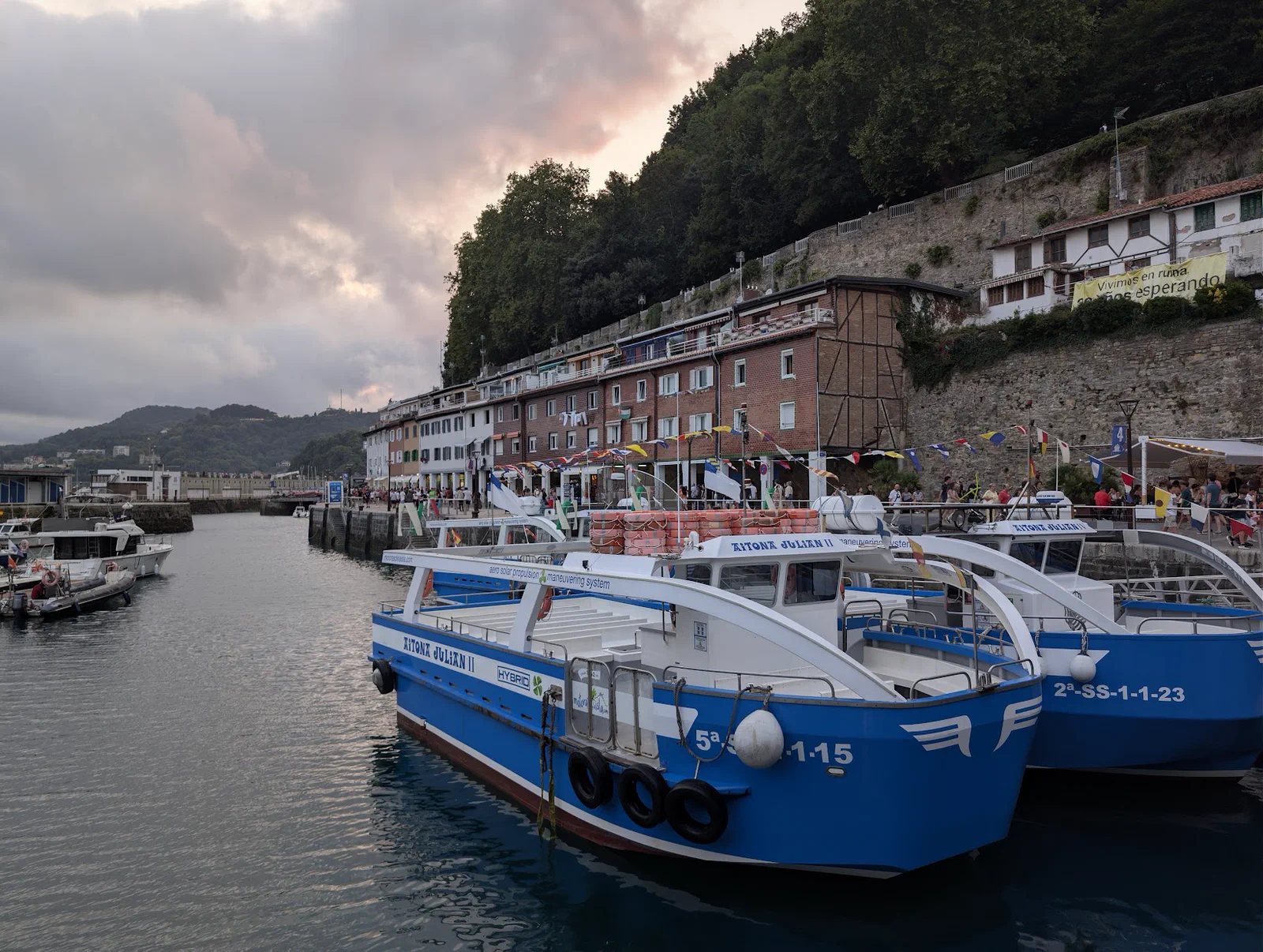 Dos motoras azules de Motoras de la Isla amarradas en el puerto de Donostia al atardecer, con las casas del muelle, las banderolas de fiestas y la ladera de Urgull al fondo