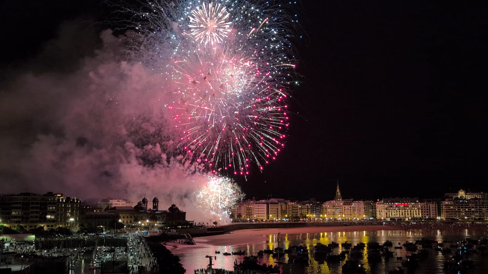 Racimo de fuegos artificiales rosas y blancos en el tramo final de la tanda, con la bahía de la Concha y los barcos fondeados debajo