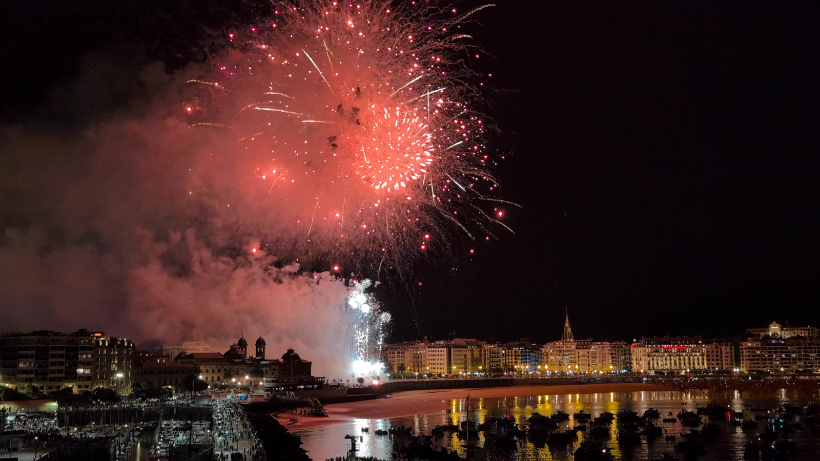 Estallido rojo de la traca final de Pirotecnia Valenciana sobre Donostia, con el humo iluminado y la playa de la Concha teñida de rojo