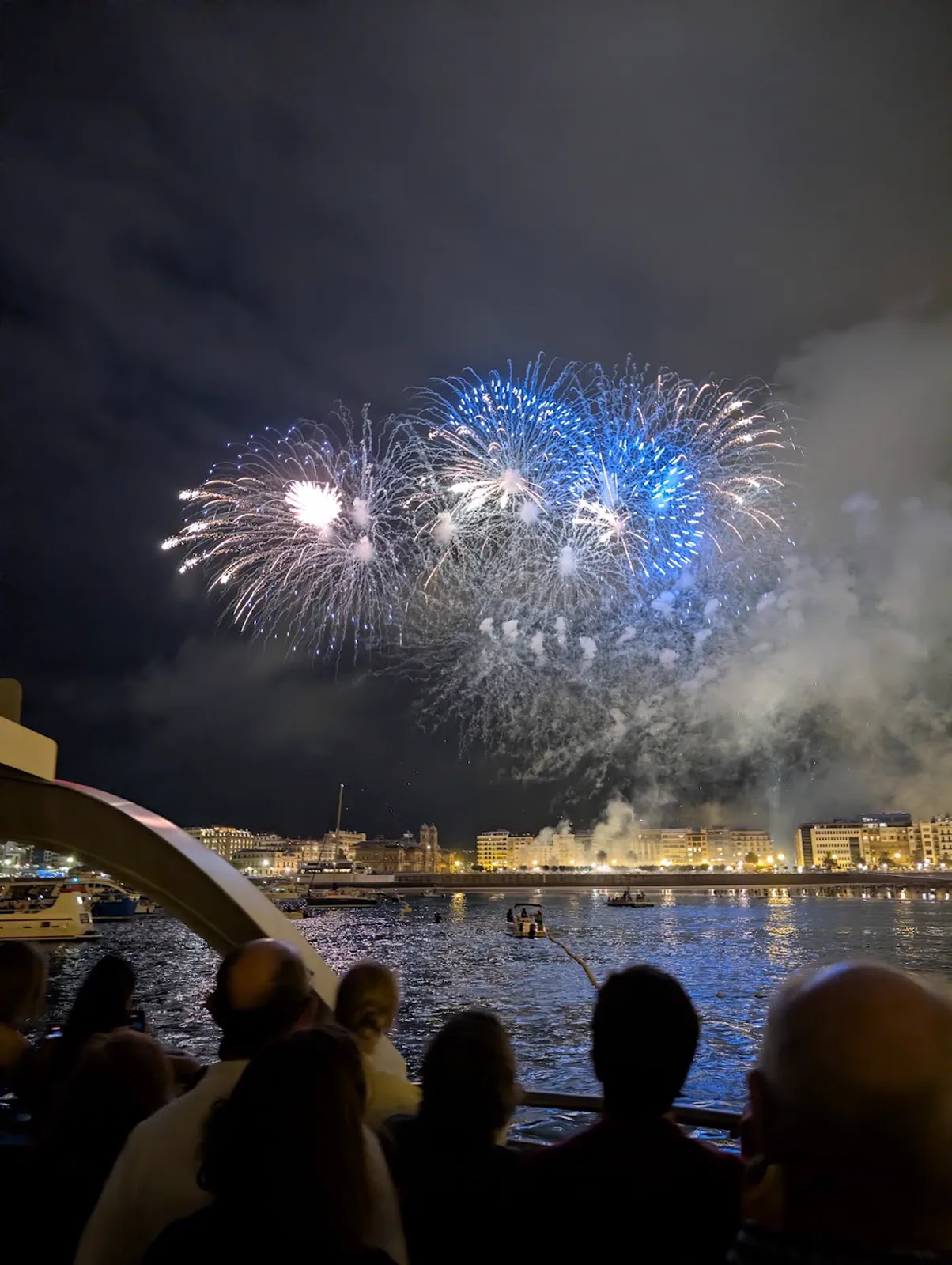 Fuegos artificiales blancos y azules abriéndose sobre la bahía de la Concha vistos desde la cubierta de la motora, con las siluetas de los pasajeros recortadas a contraluz en primer plano