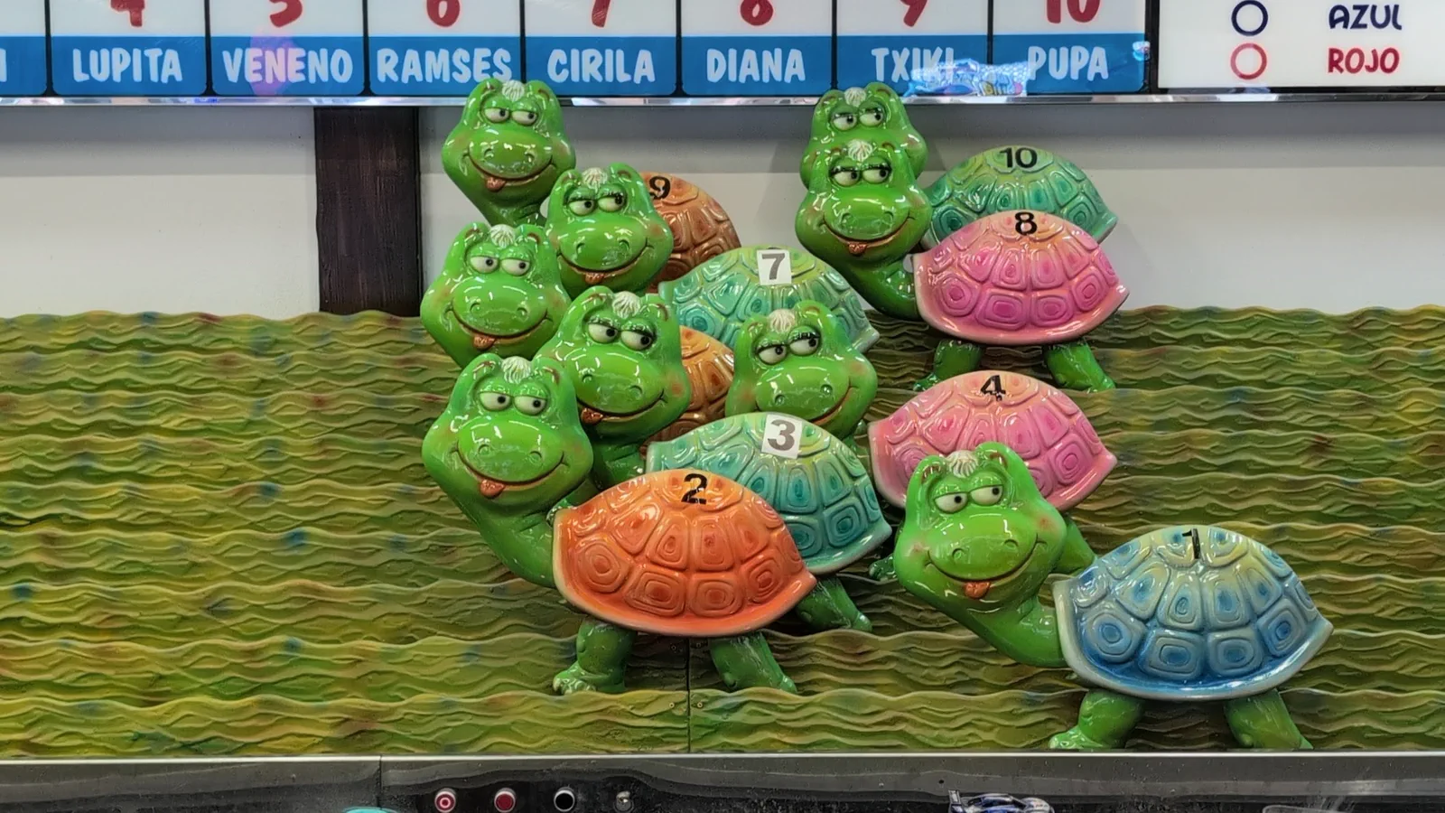 La carrera de tortugas del parque de Igueldo, con las tortugas de colores agrupadas sobre el tapete verde y el marcador con sus nombres: Lupita, Veneno, Ramsés, Cirila, Diana, Txiki y Pupa