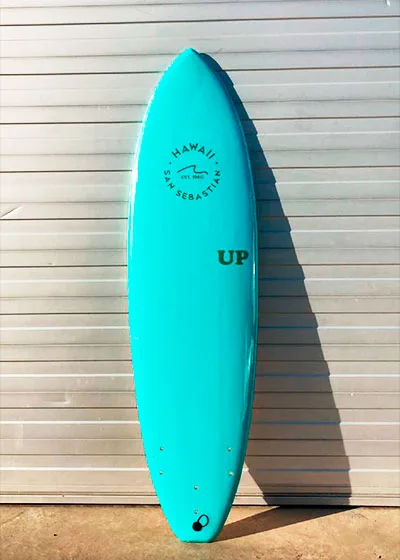 Tabla de surf softboard turquesa de alquiler con el sello «Hawaii San Sebastián» apoyada en la pared