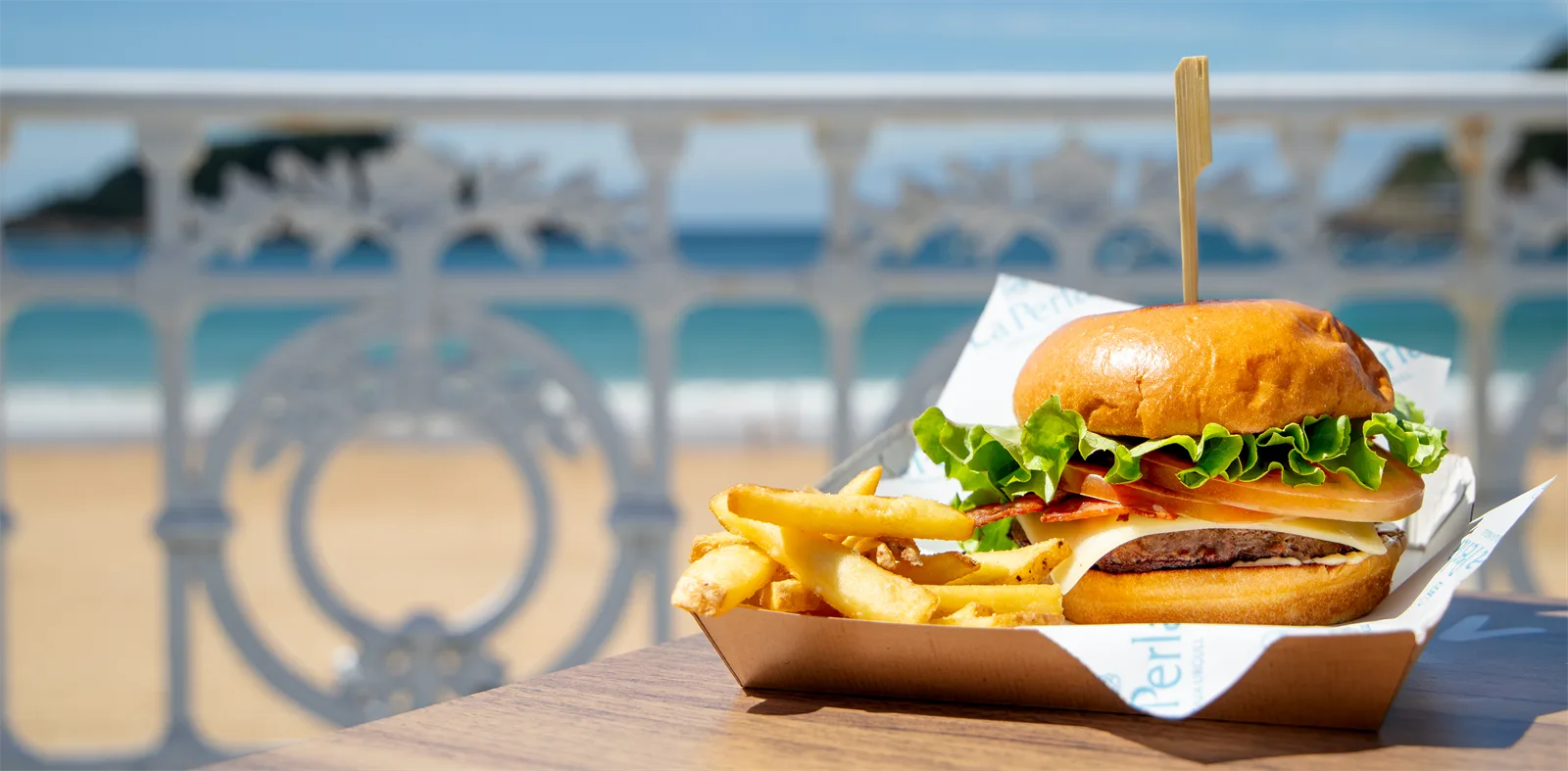 Hamburguesa con patatas de la terraza Urgull de La Perla sobre una mesa, con la barandilla de la Concha y la playa detrás