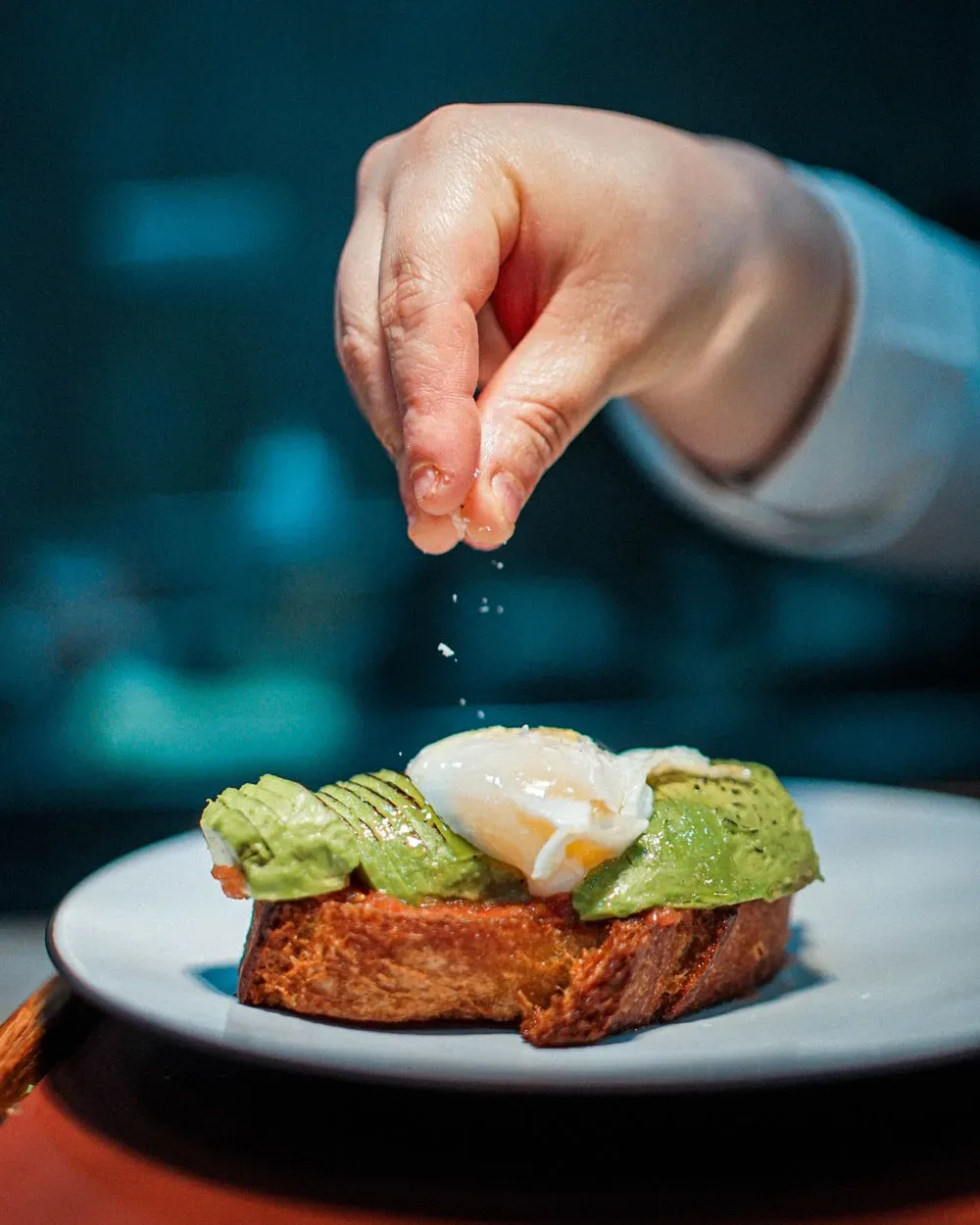 Tostada de aguacate con huevo poché a la que una mano añade una pizca de sal en Narru