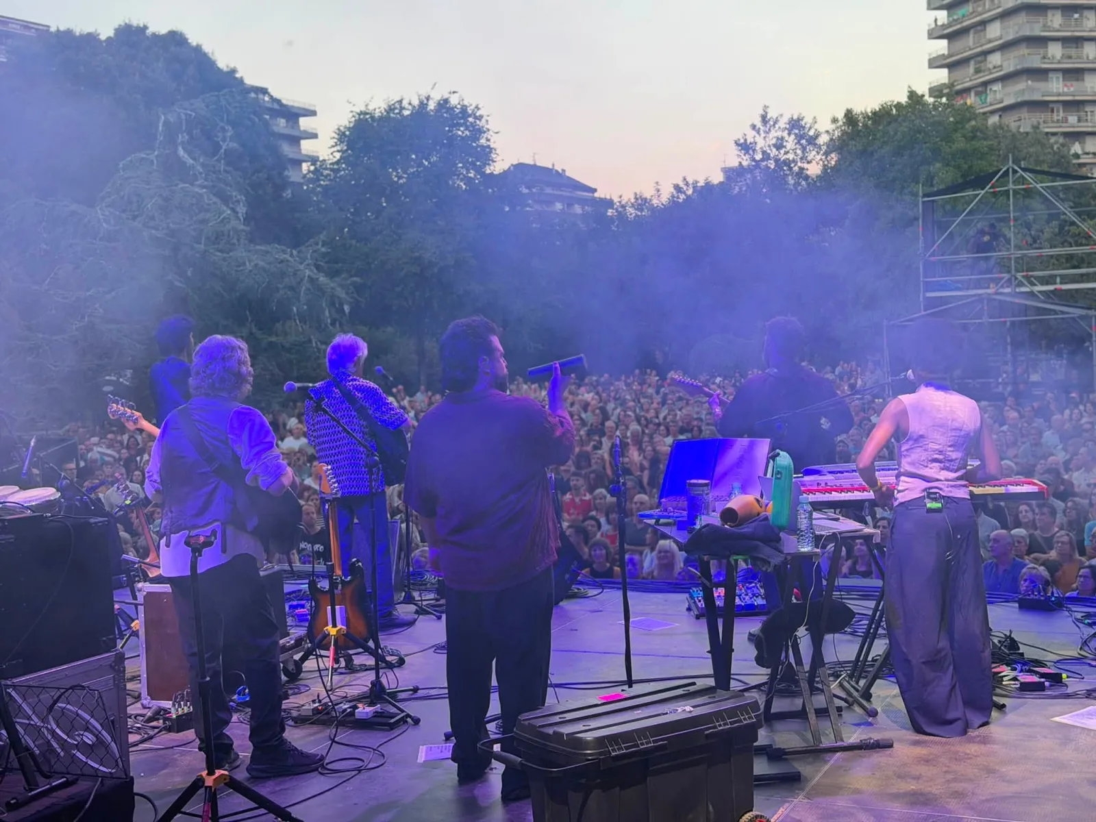 Fotografía tomada desde el escenario, de espaldas a los músicos: seis integrantes de la banda en primer término y, ante ellos, la explanada de la Alameda Miramar cubierta de público bajo un cielo de atardecer