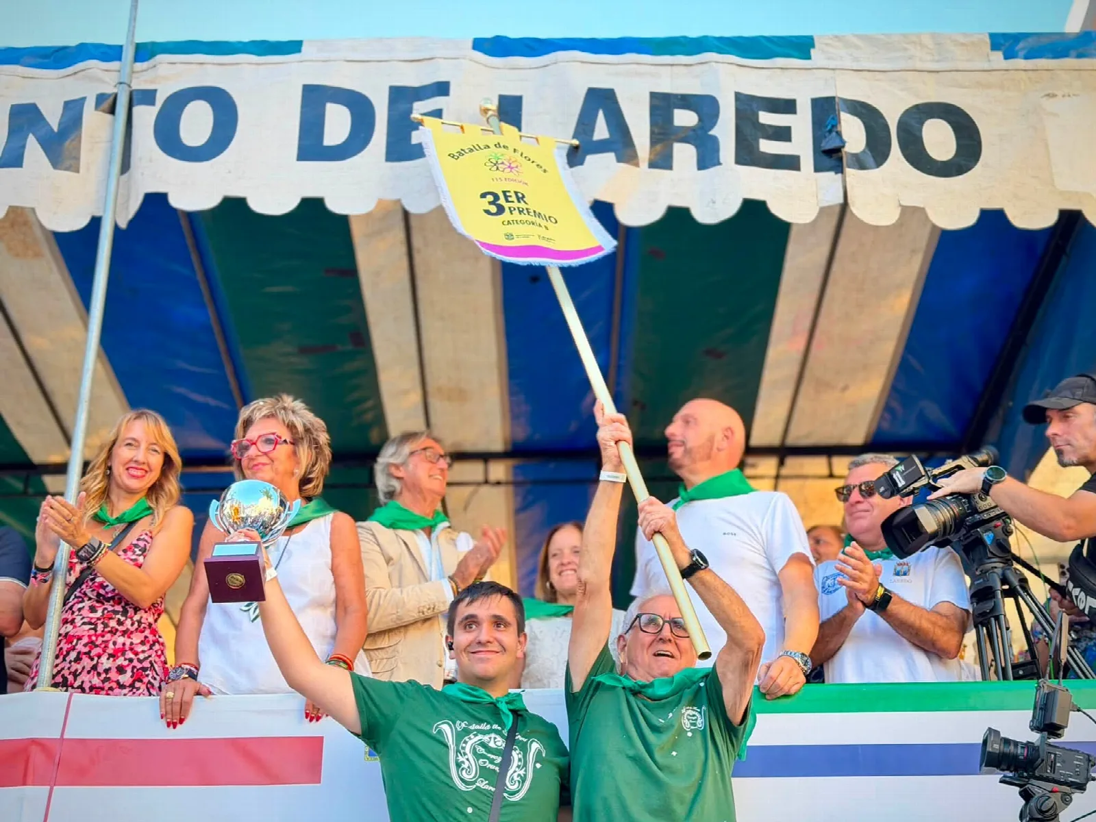 Dos personas con camiseta verde levantan ante la tribuna un trofeo y un banderín amarillo con la leyenda «3er premio categoría B», mientras las autoridades aplauden