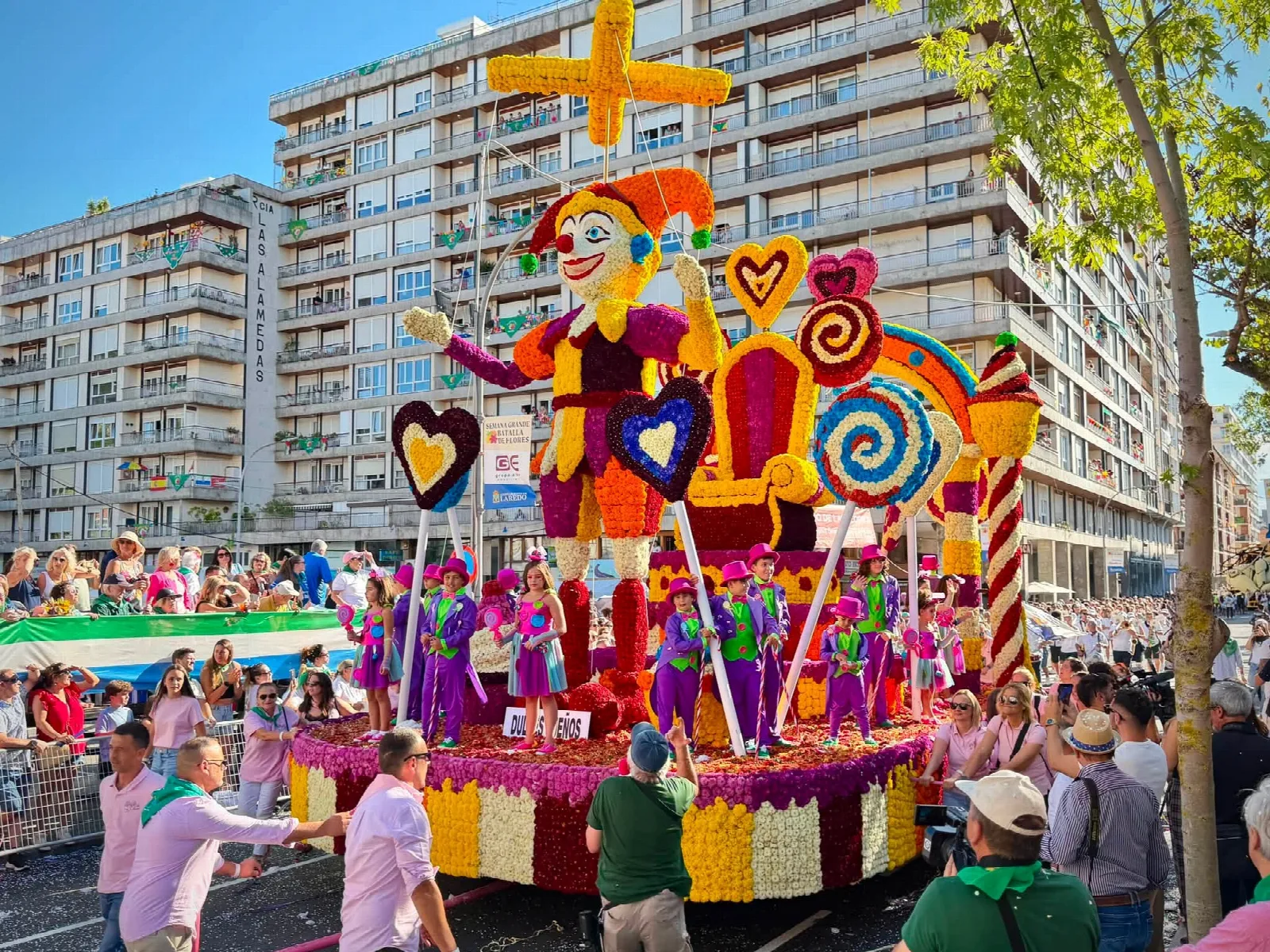 Carroza de flor con figuras de caramelos y piruletas gigantes y una marioneta de arlequín, con la placa del título delante