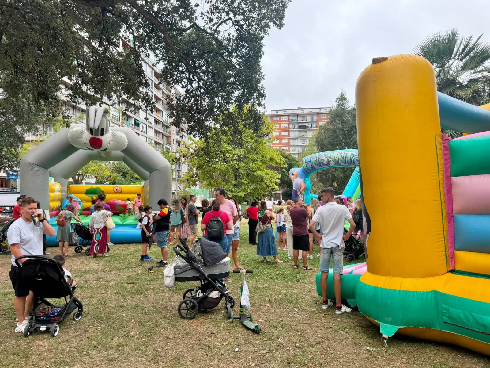 Dos castillos hinchables en un parque: a la izquierda uno con un arco en forma de conejo de dibujos animados y niños saltando dentro, a la derecha otro de rayas amarillas, rosas y verdes, con familias y carritos de bebé esperando alrededor