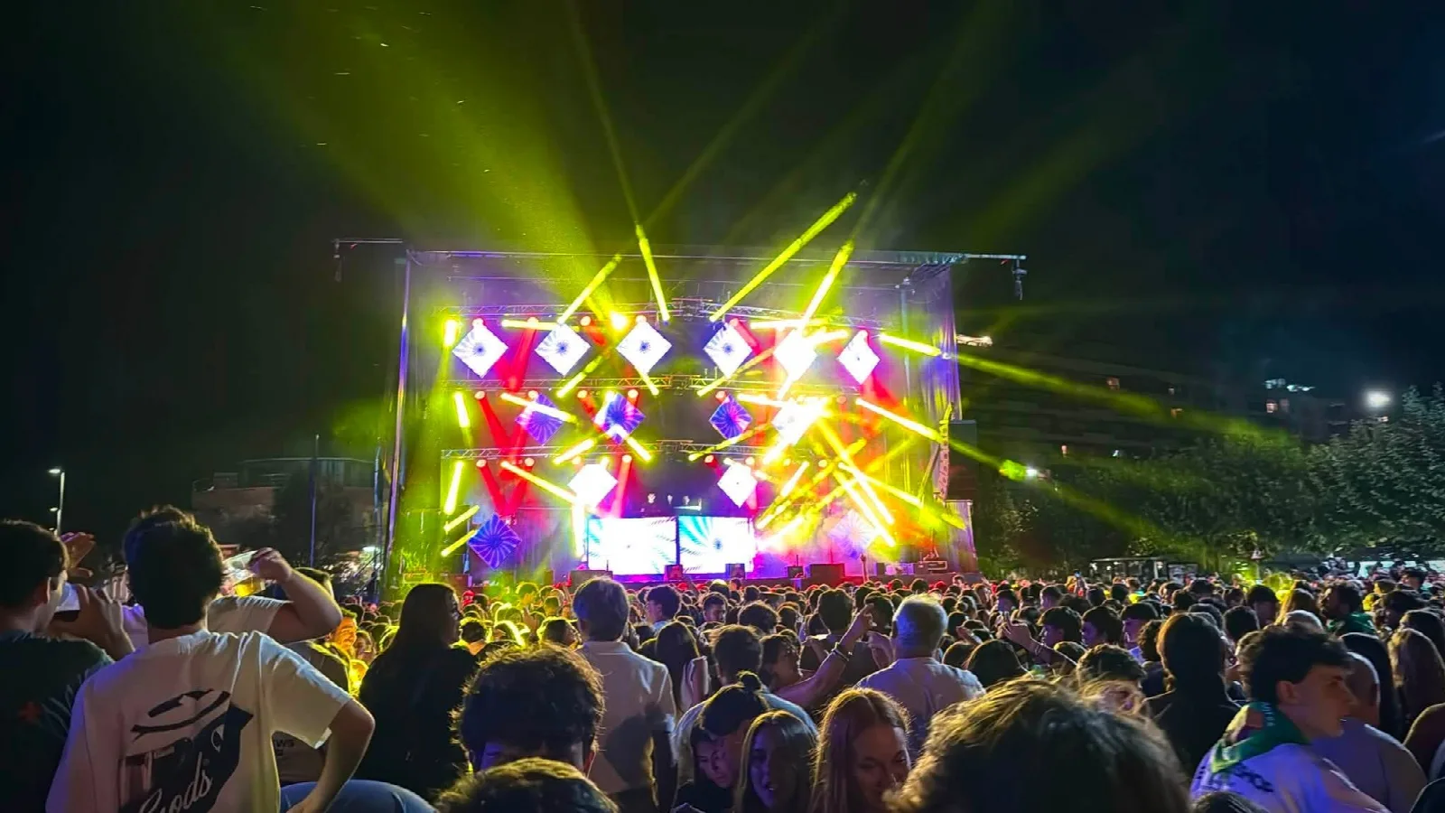 Escenario de gran formato con pantallas y decenas de haces de láser amarillos y rojos cruzándose sobre una multitud de gente joven en un parque de noche