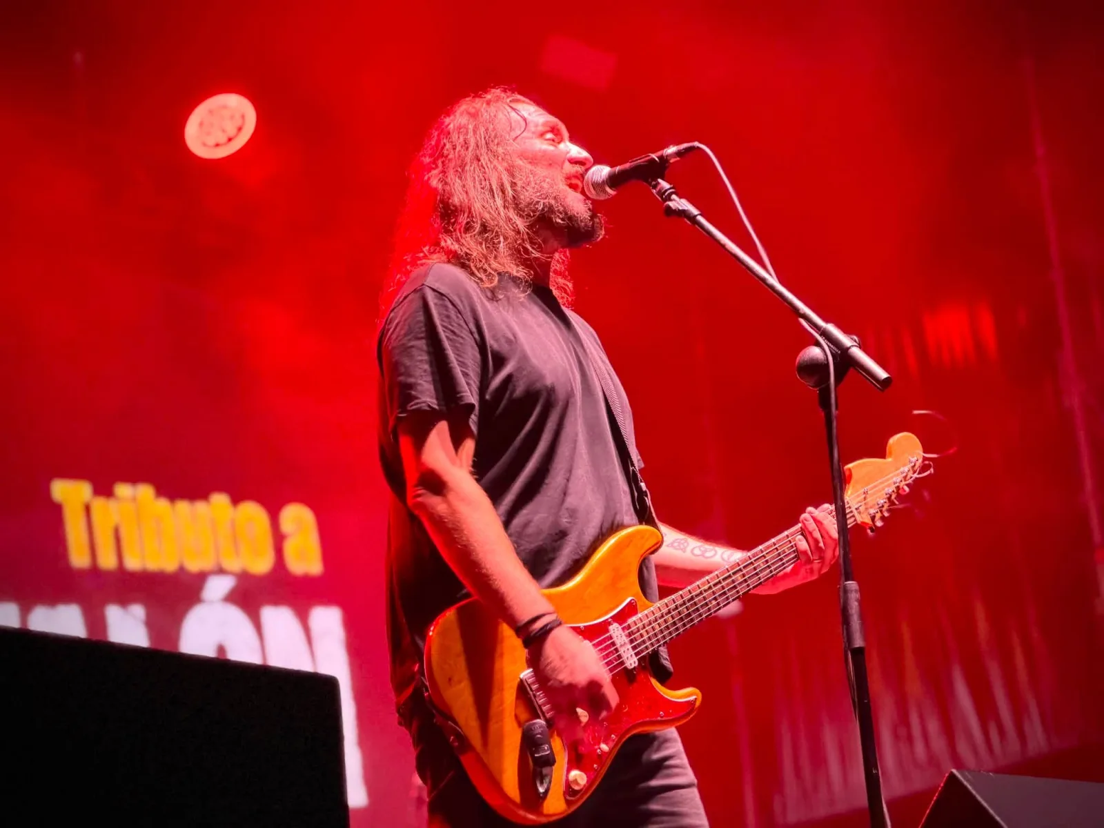 Un guitarrista de melena y barba canta al micrófono con una guitarra naranja, iluminado en rojo, ante un photocall en el que se lee «Tributo a»