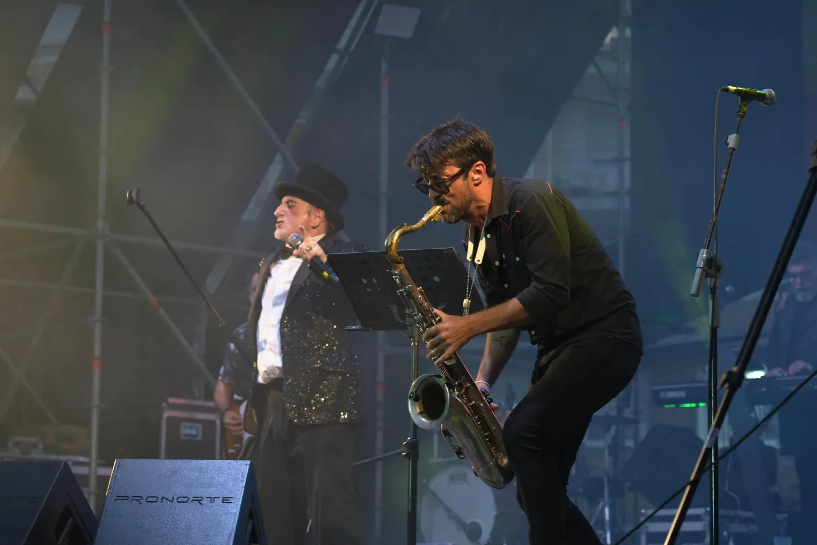 Un saxofonista tocando inclinado hacia delante en primer plano y, detrás, el cantante de la Orquesta Mondragón con chistera y chaqueta de lentejuelas ante un micrófono