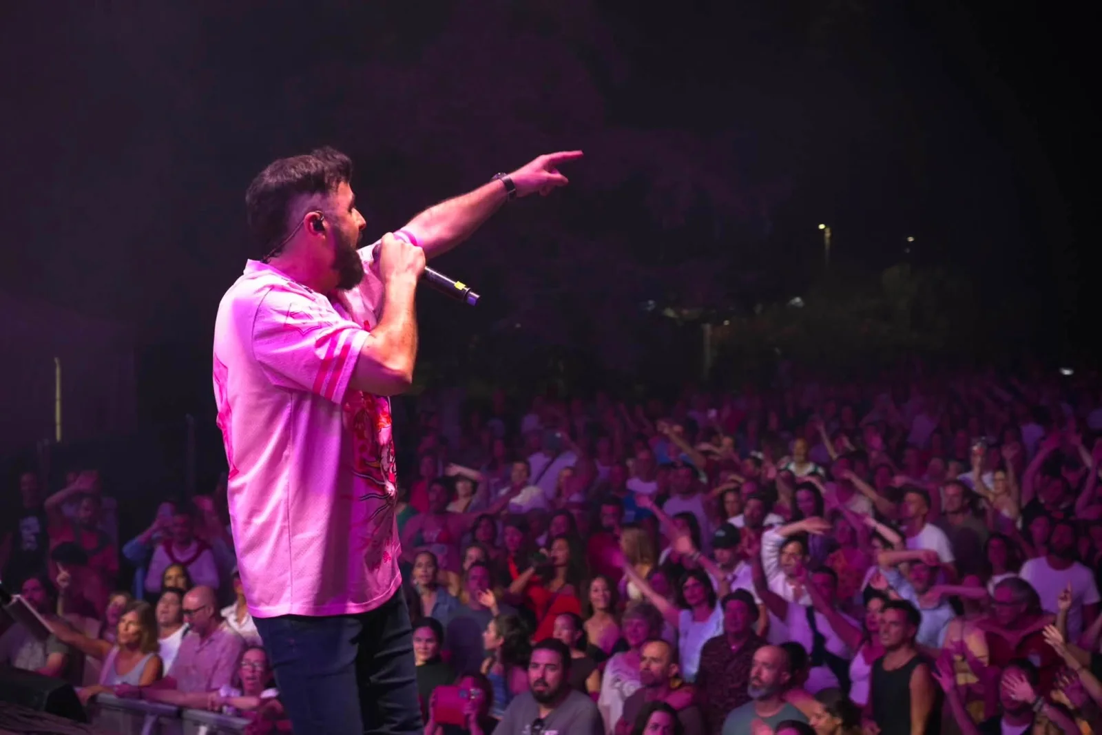 Huecco de perfil en el borde del escenario, cantando con el micrófono en una mano y señalando al público con la otra, bajo una luz rosa intensa que ilumina a cientos de personas con los brazos en alto