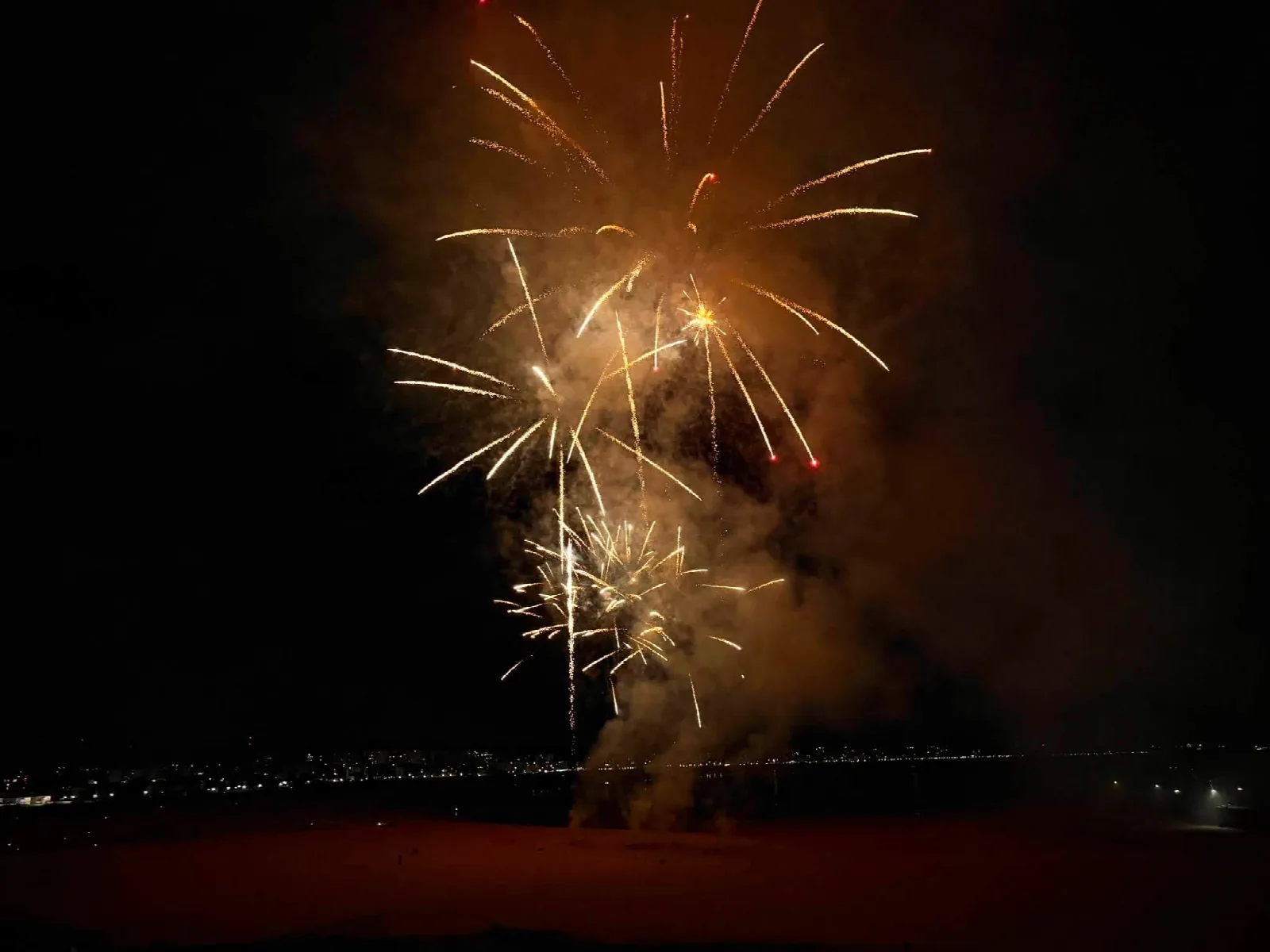 Varias explosiones de fuegos artificiales doradas encadenadas entre una densa nube de humo iluminada, sobre la playa a oscuras
