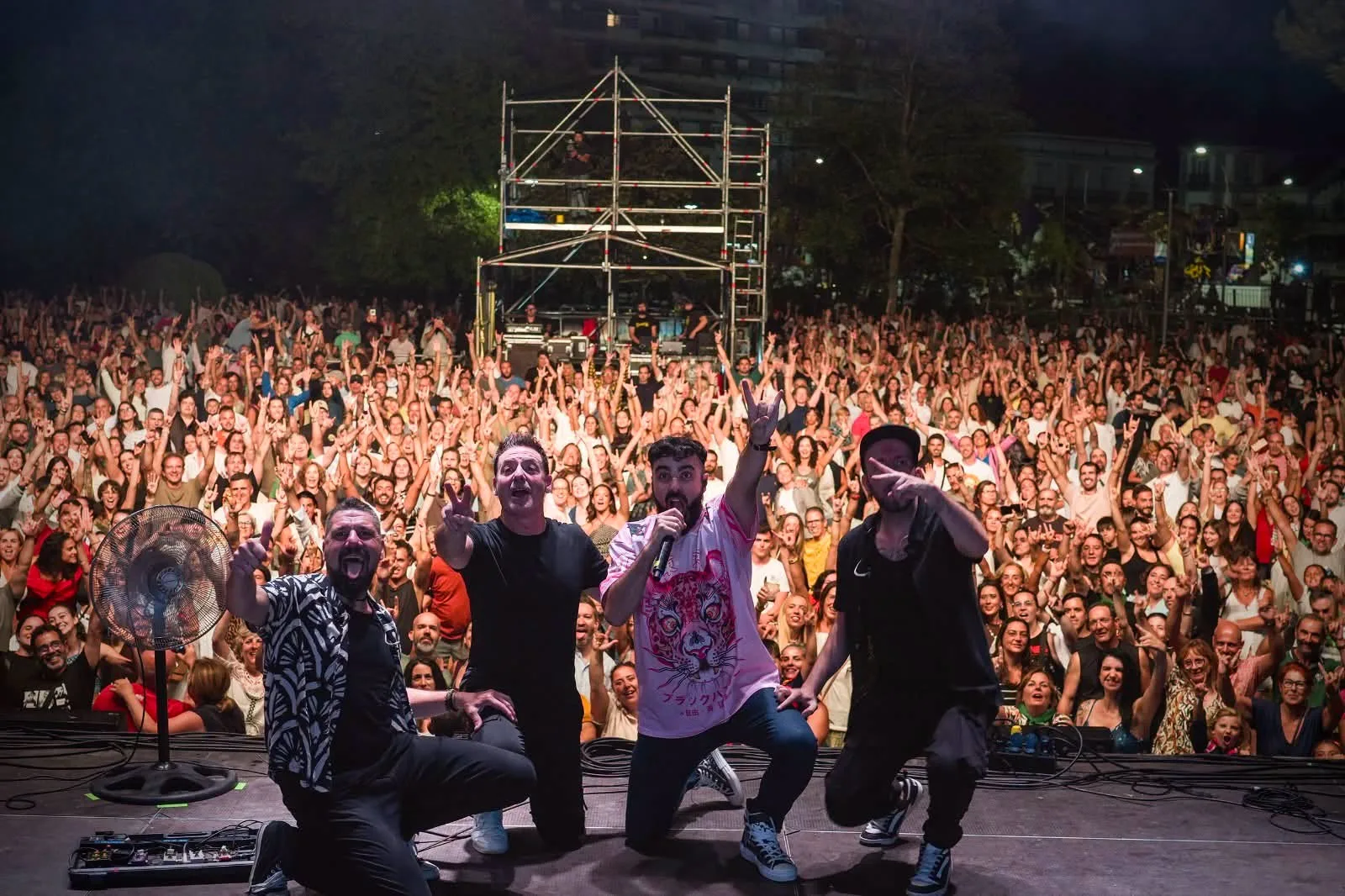 Cuatro músicos posando agachados en el borde del escenario con gestos de celebración y, detrás de ellos, cientos de personas con los brazos levantados llenando la explanada de la Alameda Miramar de noche
