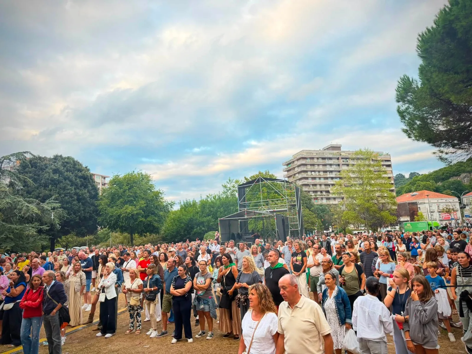 Plano frontal de varios cientos de personas de pie en la explanada de la Alameda Miramar, todavía de día y con el cielo nublado, con la torre de control de sonido montada en mitad del público y los bloques de viviendas de Laredo al fondo