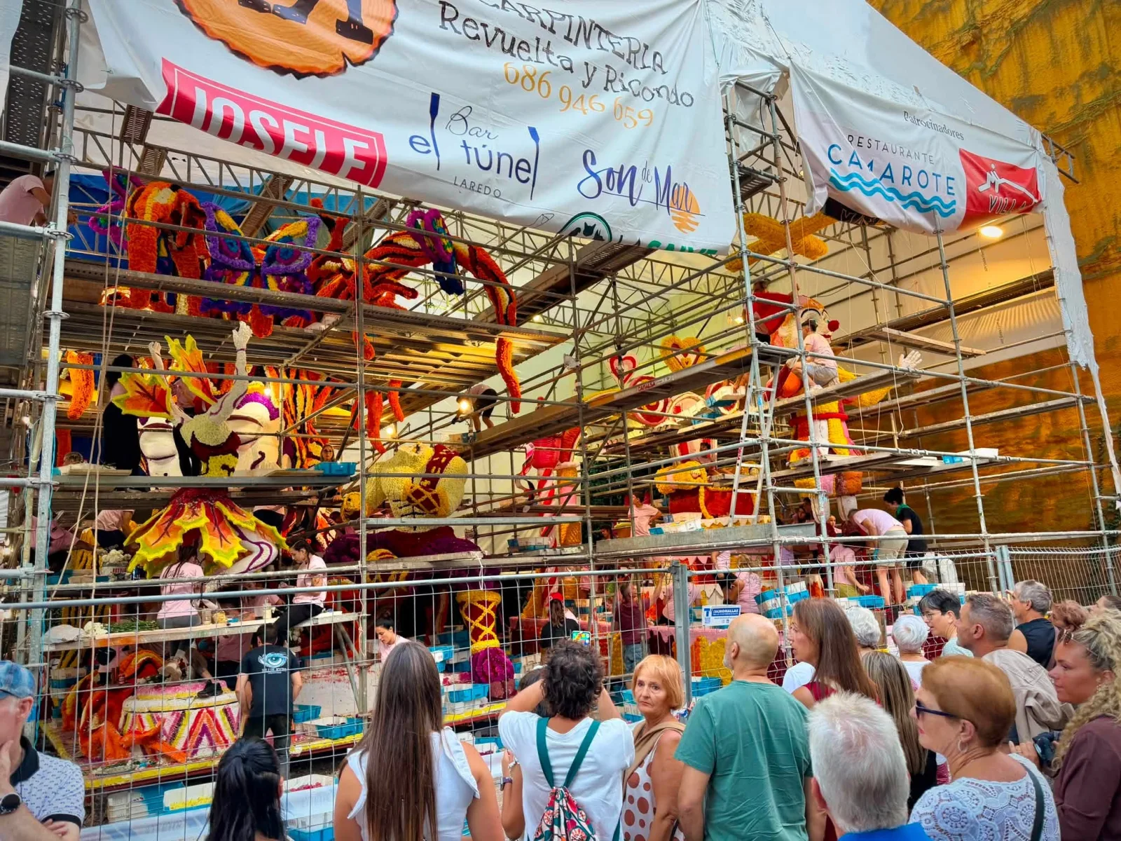 Grupo de personas apoyadas en una valla metálica mirando hacia el interior de una carpa de montaje donde varias personas trabajan sobre una carroza