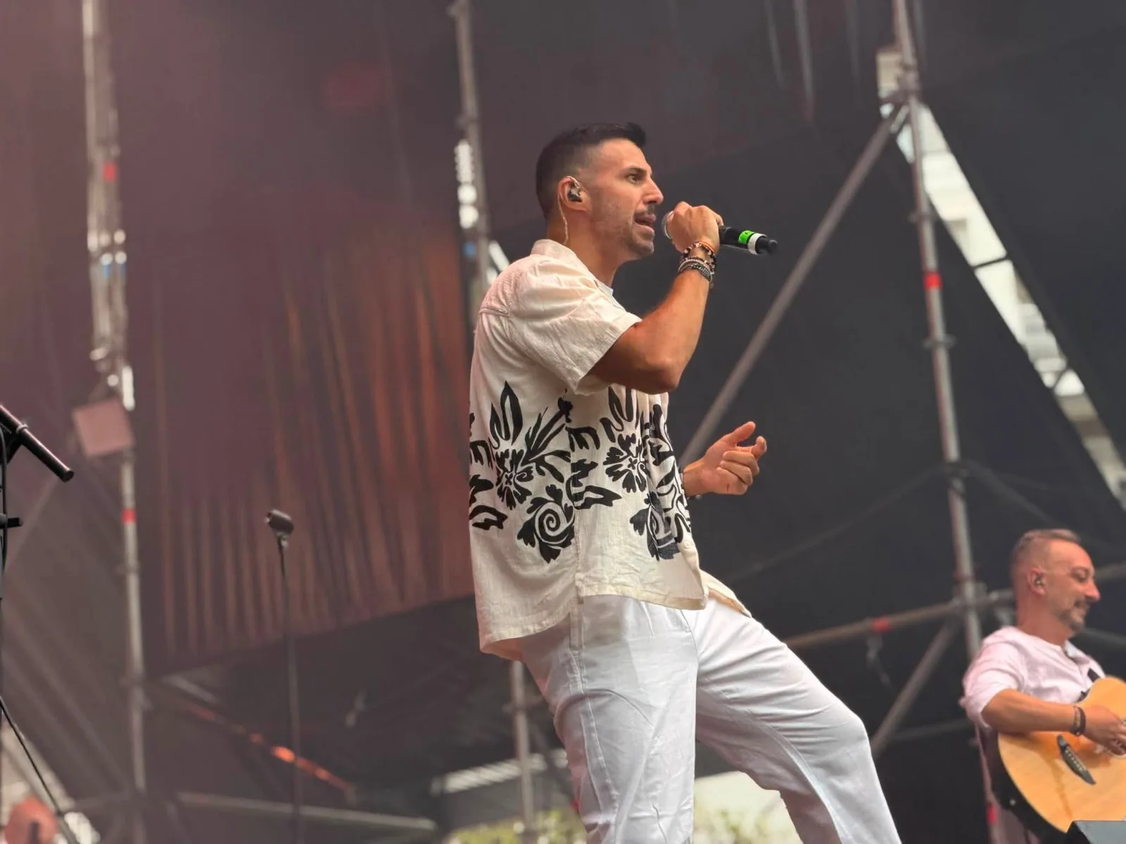 Primer plano del cantante del tributo, de perfil y con el micrófono en la mano, vestido con camisa blanca de estampado floral negro y pantalón blanco, con un guitarrista acústico desenfocado a la derecha