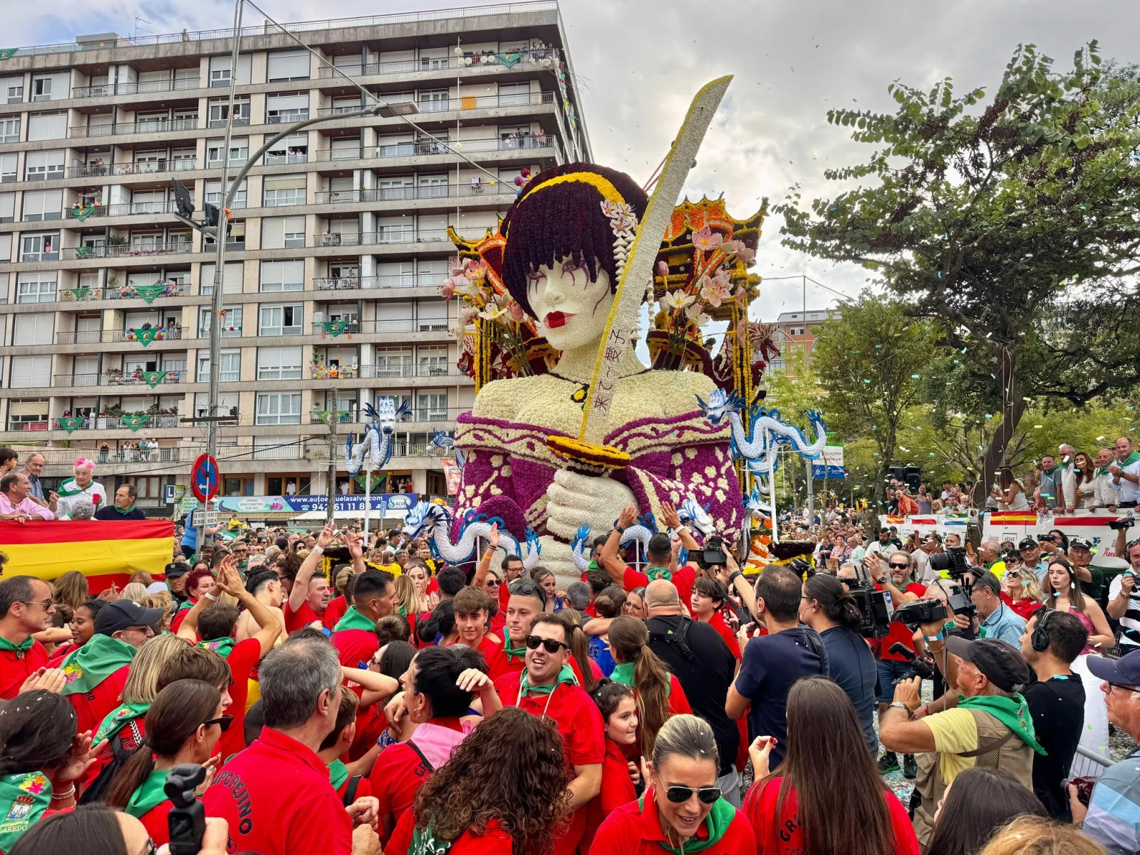La carroza «Aiko», un busto femenino de flor blanca con kimono y catana, rodeada por los carrocistas del Grupo Pejino vestidos de rojo